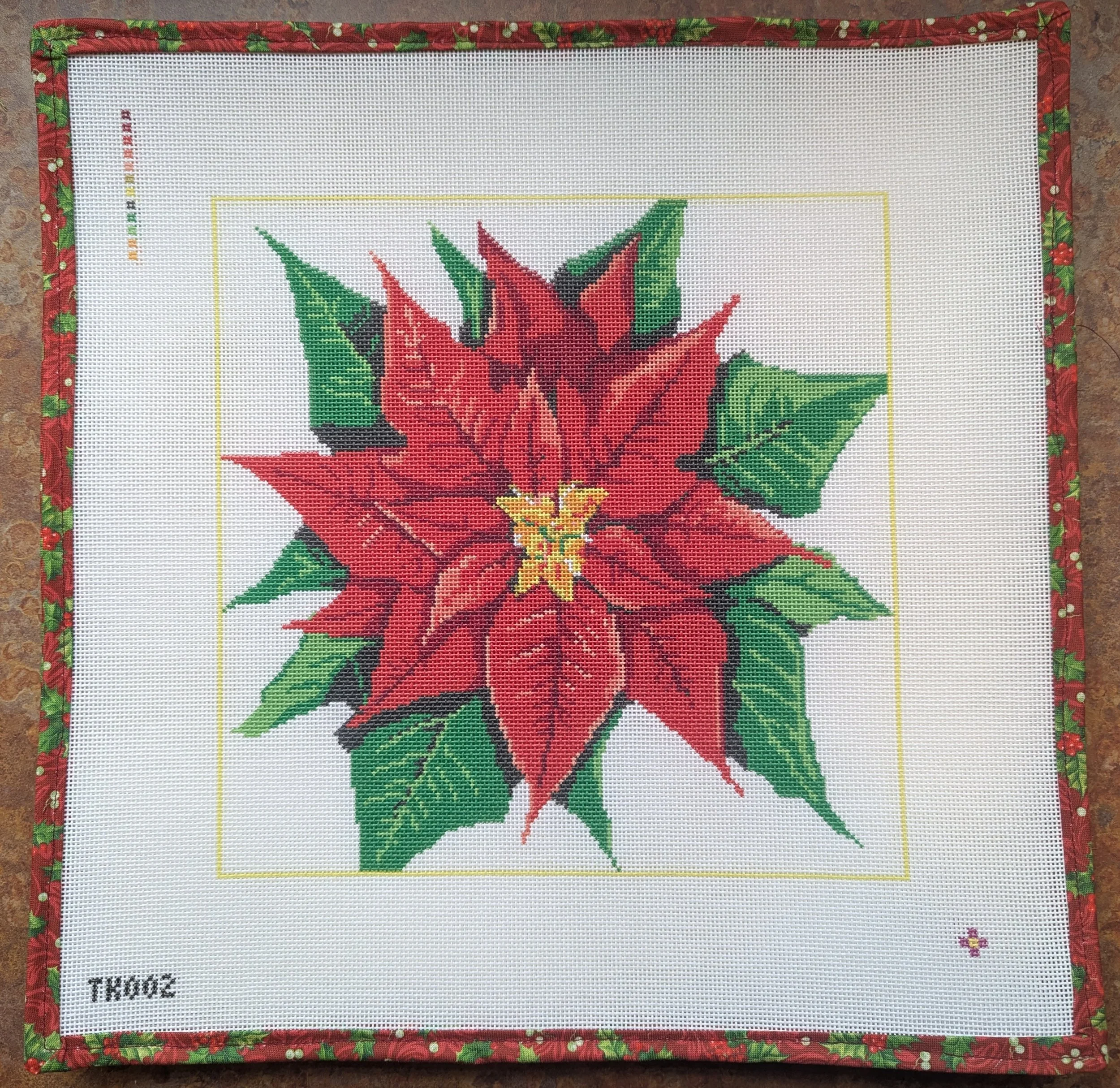 A cross-stitch embroidery kit featuring a poinsettia flower with red petals, green leaves, and a yellow center, framed with a festive red and green holly border.