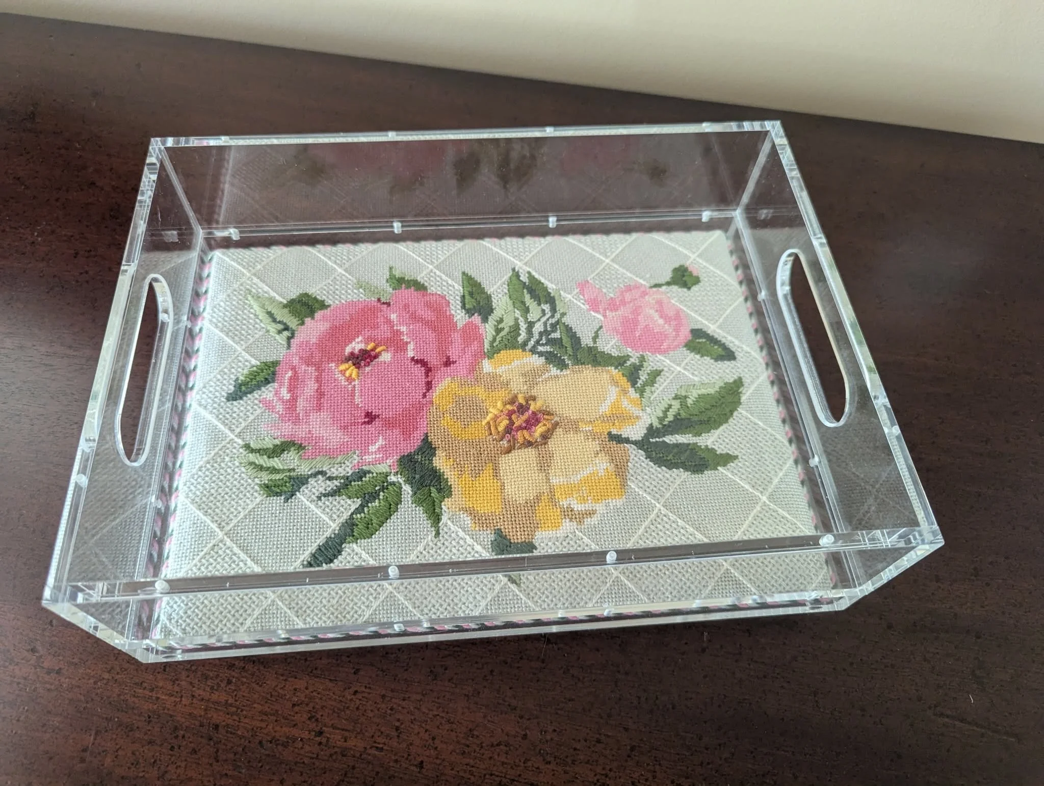 A clear acrylic box on a dark wooden table, with a cross-stitch embroidery of pink, yellow, and green flowers inside.