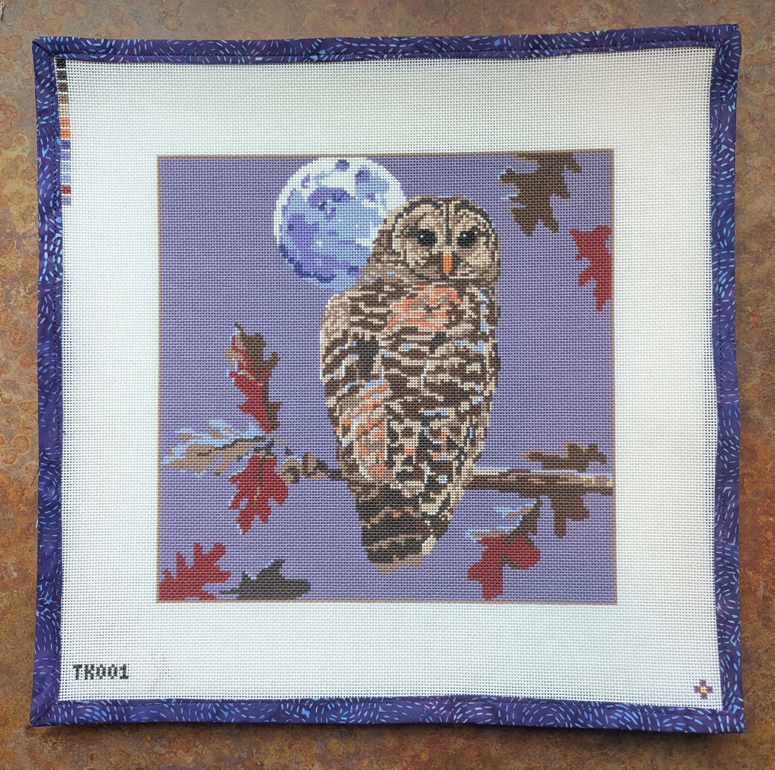 Cross-stitch embroidery artwork featuring an owl, a night sky with a moon and planet, surrounded by autumn leaves and flowers, bordered with a blue patterned fabric.