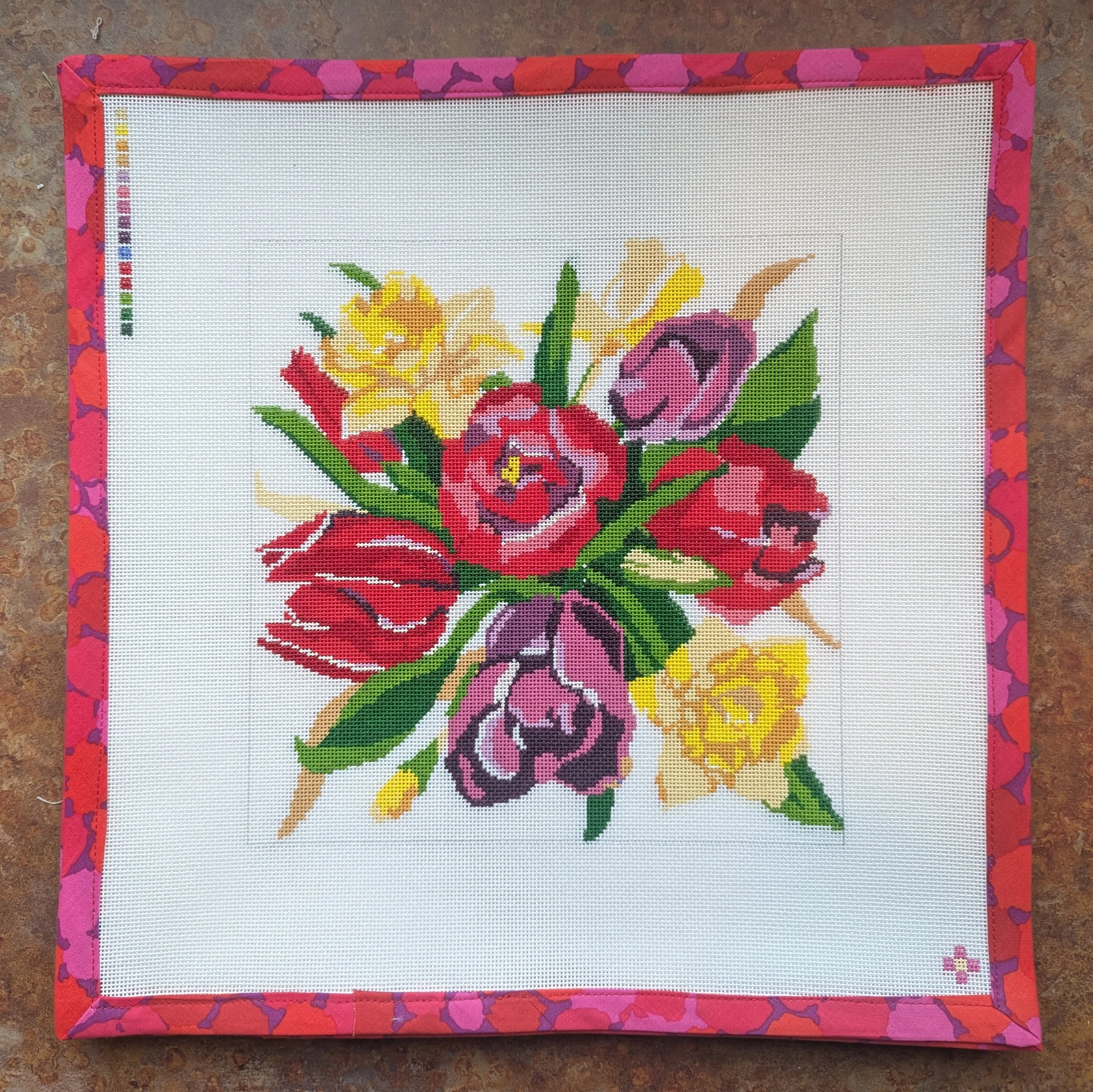A framed floral cross-stitch embroidery with colorful flowers and green leaves, surrounded by a pink and red patterned fabric border.