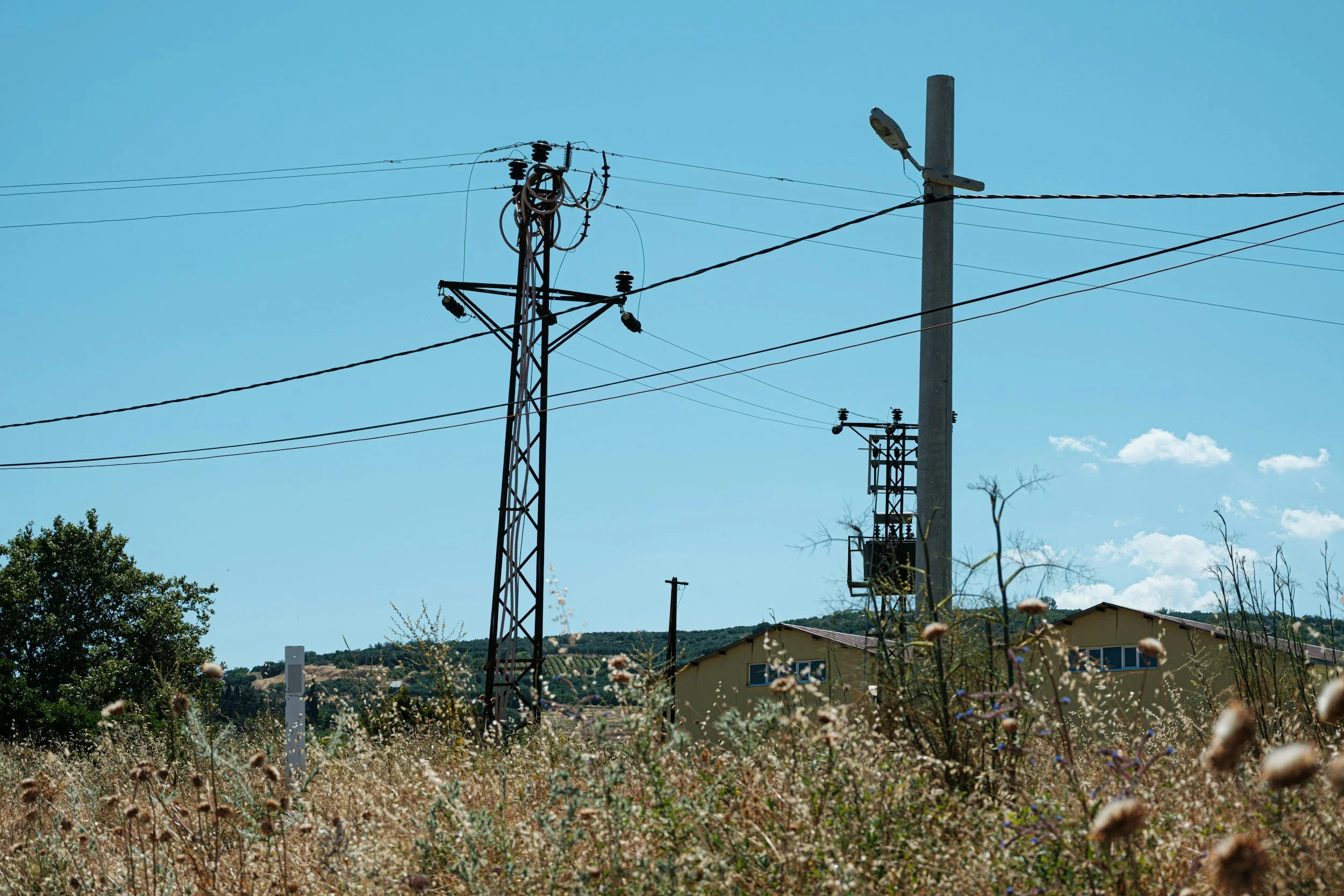 A clear blue sky with electrical power line poles and wires, some trees, and a building seen in the background.