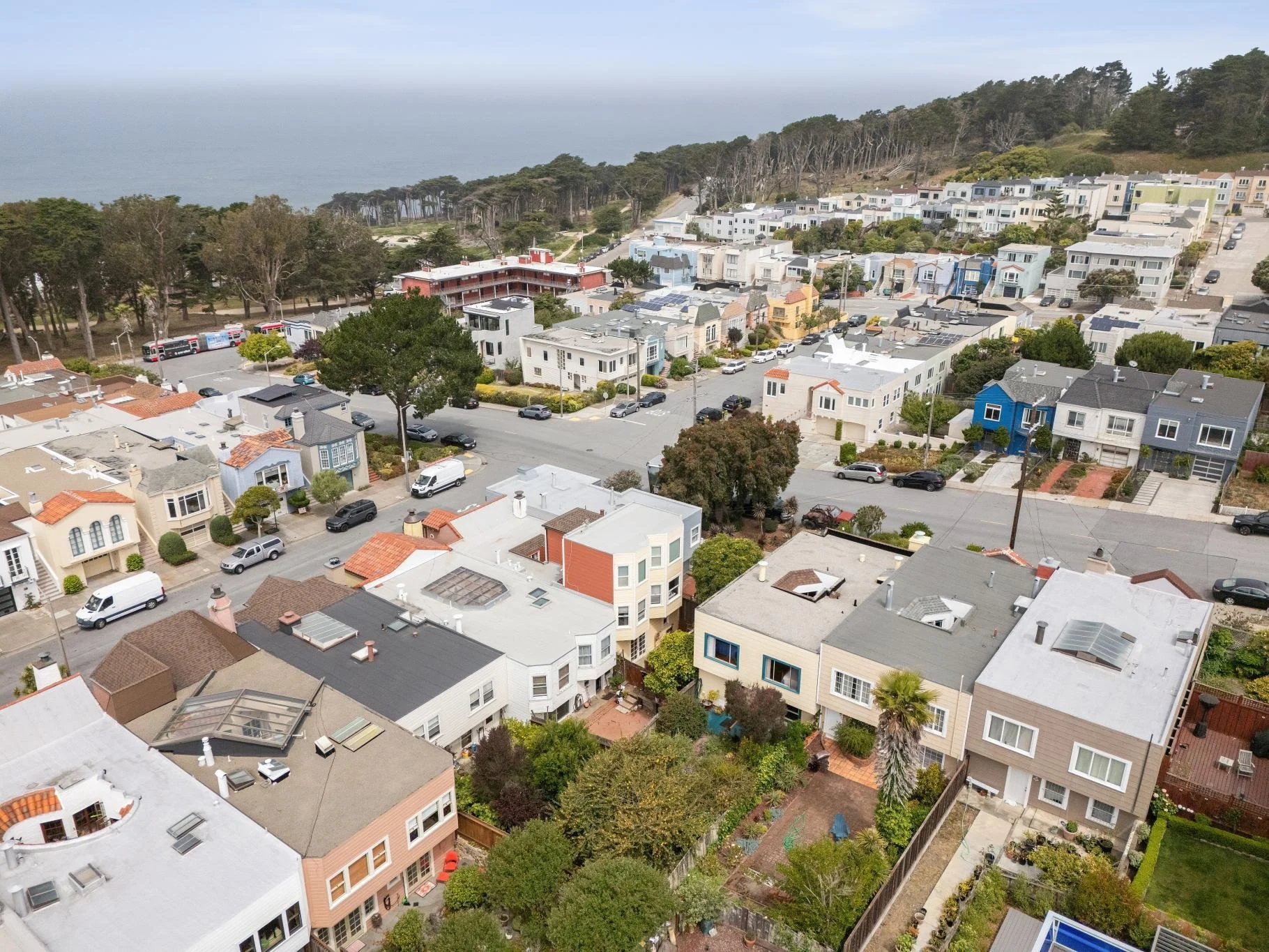 Aerial view of a coastal residential neighborhood with houses, trees, parked cars, and a roadway, near the ocean with a forested area in the background.