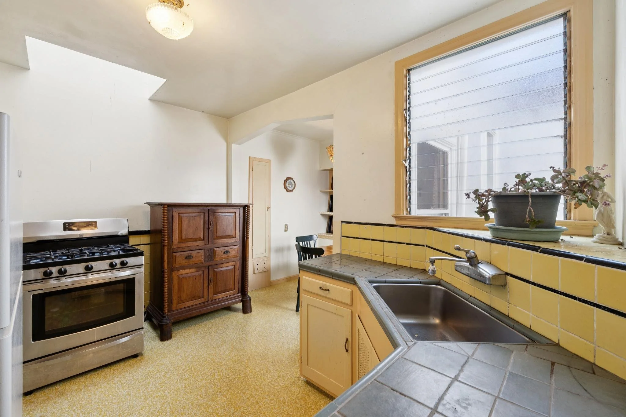 A vintage kitchen with yellow tiled counters, a stainless steel oven, a wooden cabinet, a potted plant on the sink, and a large window with a view outside.
