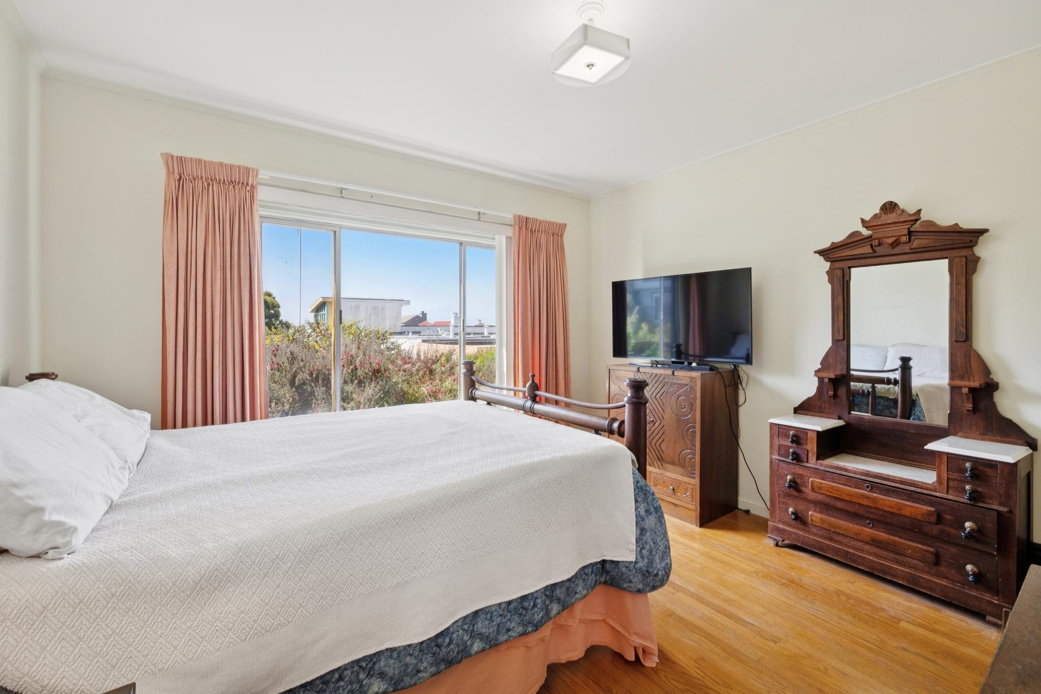 A bedroom with a large window, pink curtains, and a view of trees and rooftops outside. There is a bed with white bedding and pillows, a wooden dresser with a flat-screen TV, and an antique wooden mirror and dresser. The room has a wooden floor and w