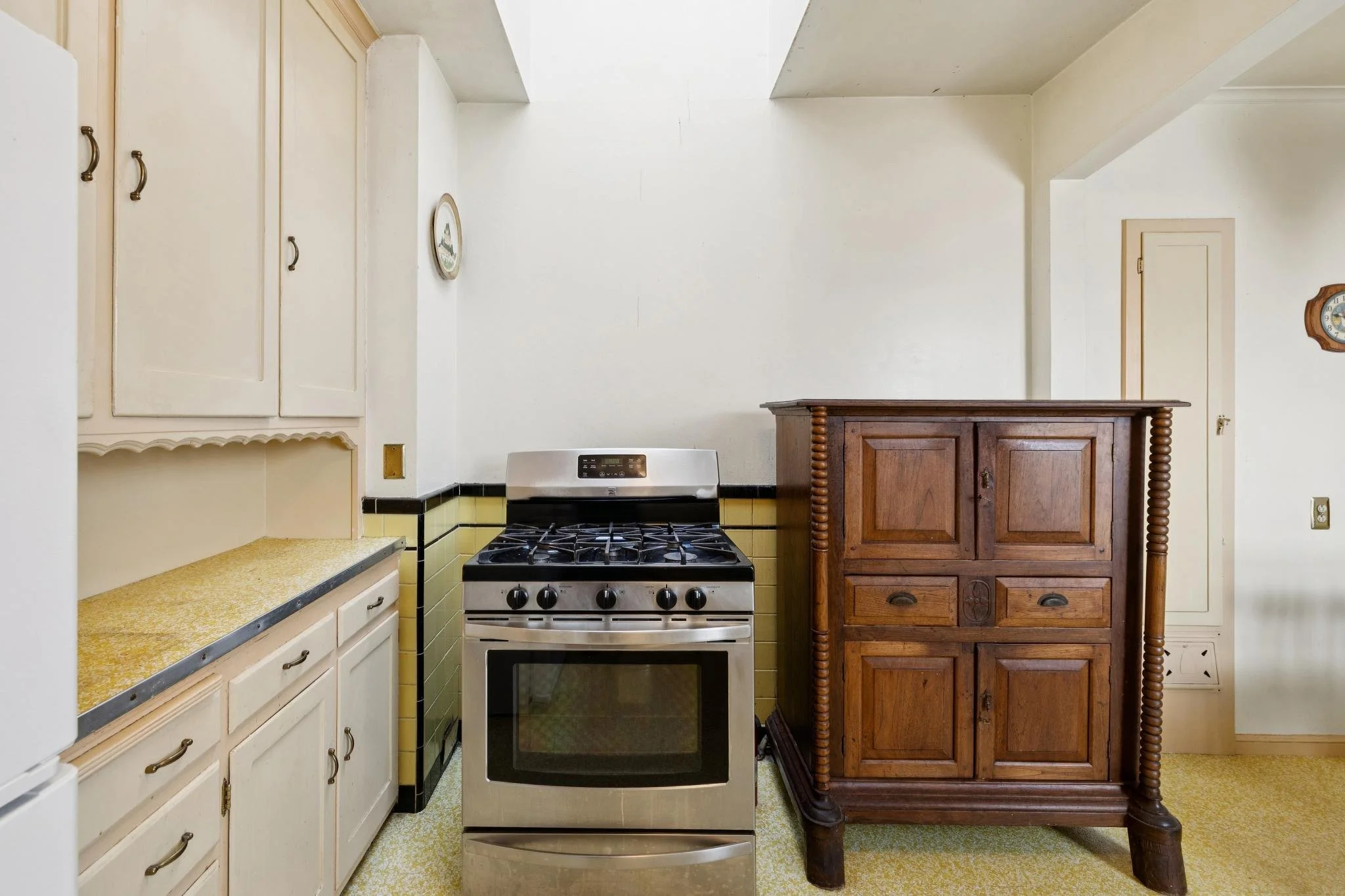 A small kitchen corner with yellow and white cabinets, a white stove, a wooden cabinet, and a wall clock.