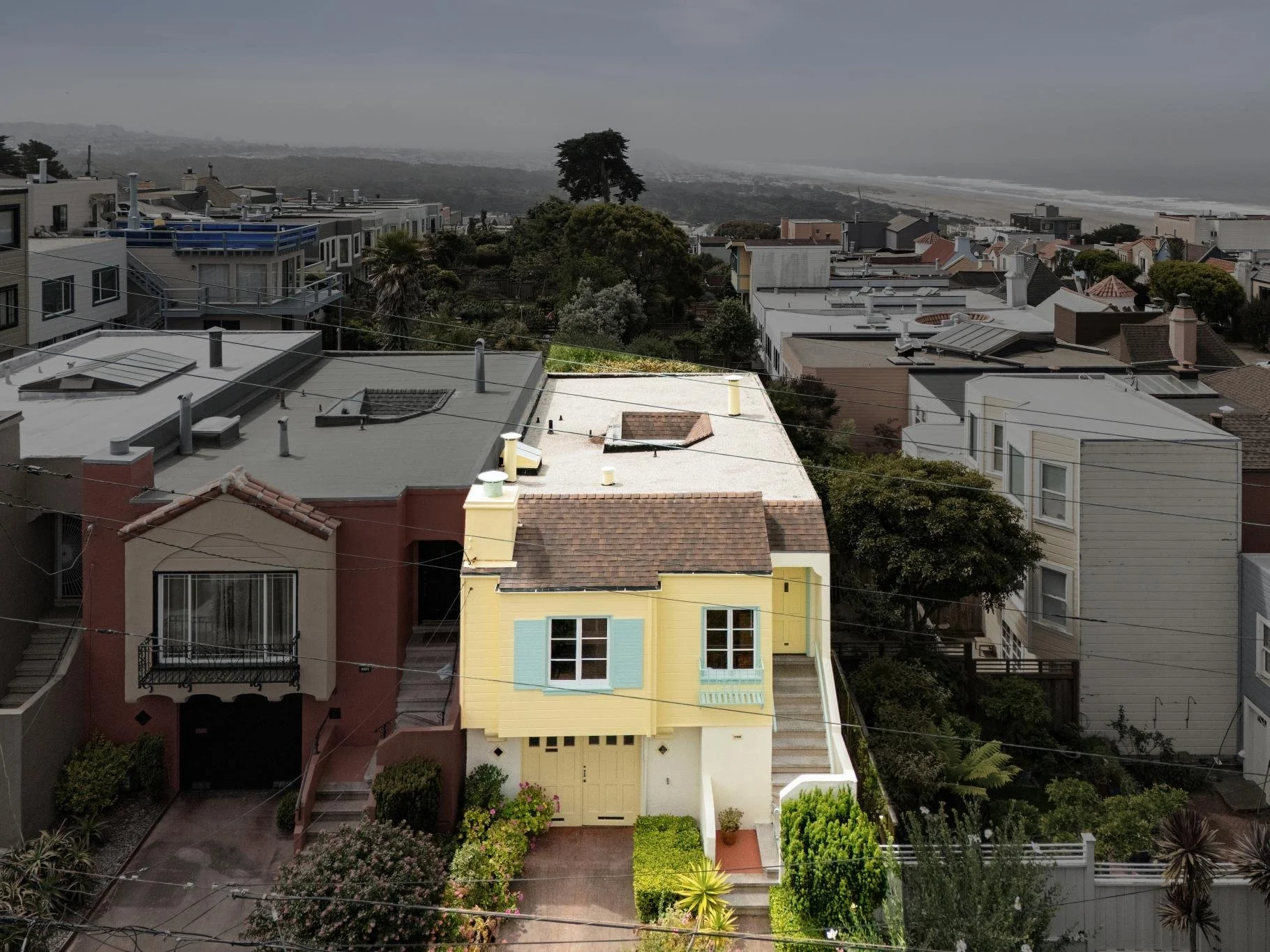 Coastal neighborhood with colorful houses, trees, and a view of the ocean in the distance under a cloudy sky.