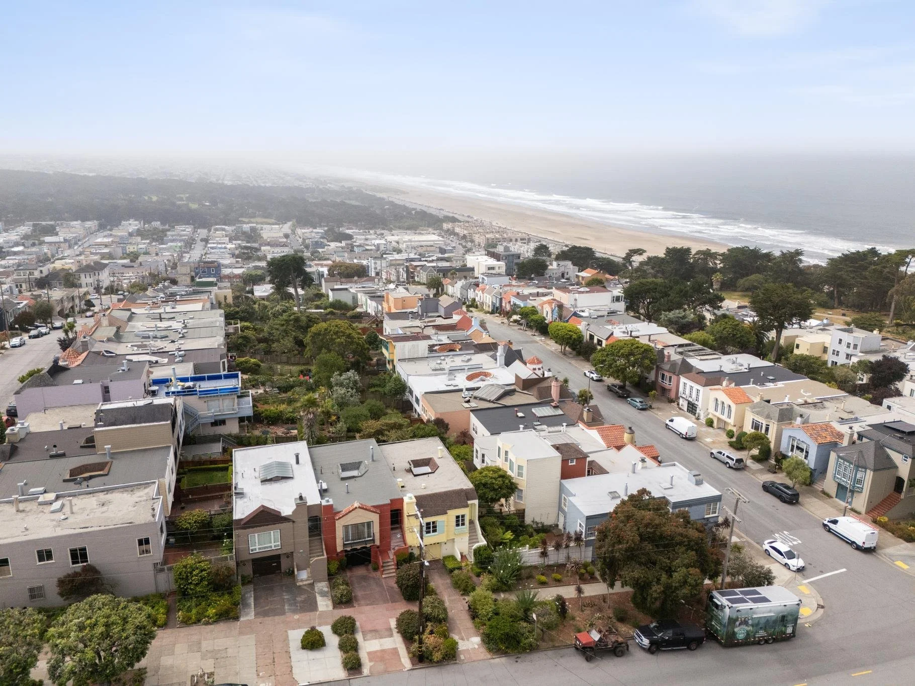 Aerial view of a coastal residential neighborhood with colorful houses, streets, trees, and a beach with waves in the background under a partly cloudy sky.