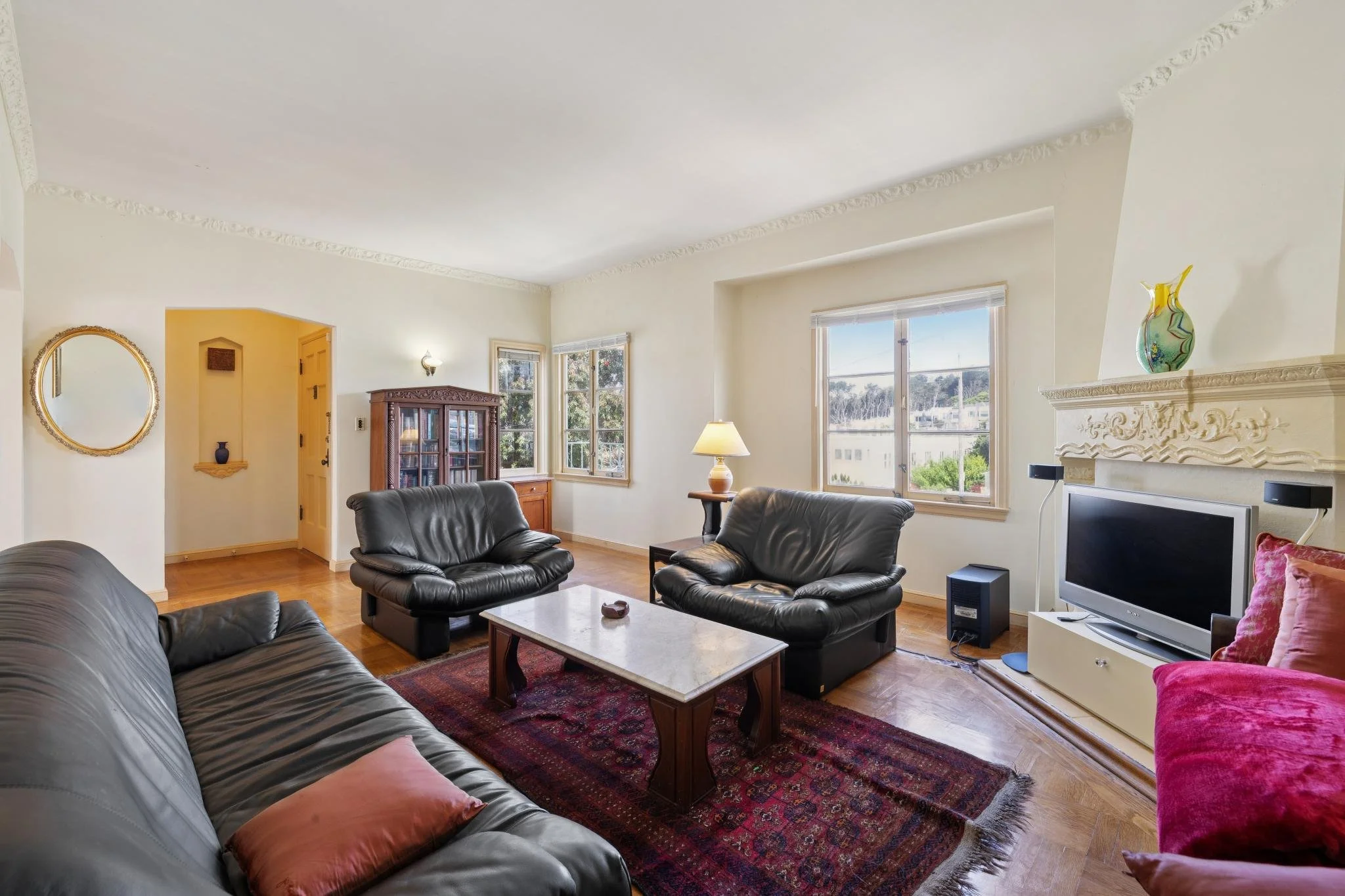 Living room with leather sofas, wooden coffee table on a red rug, fireplace with decorative molding, flat-screen TV, and large windows showing trees outside.