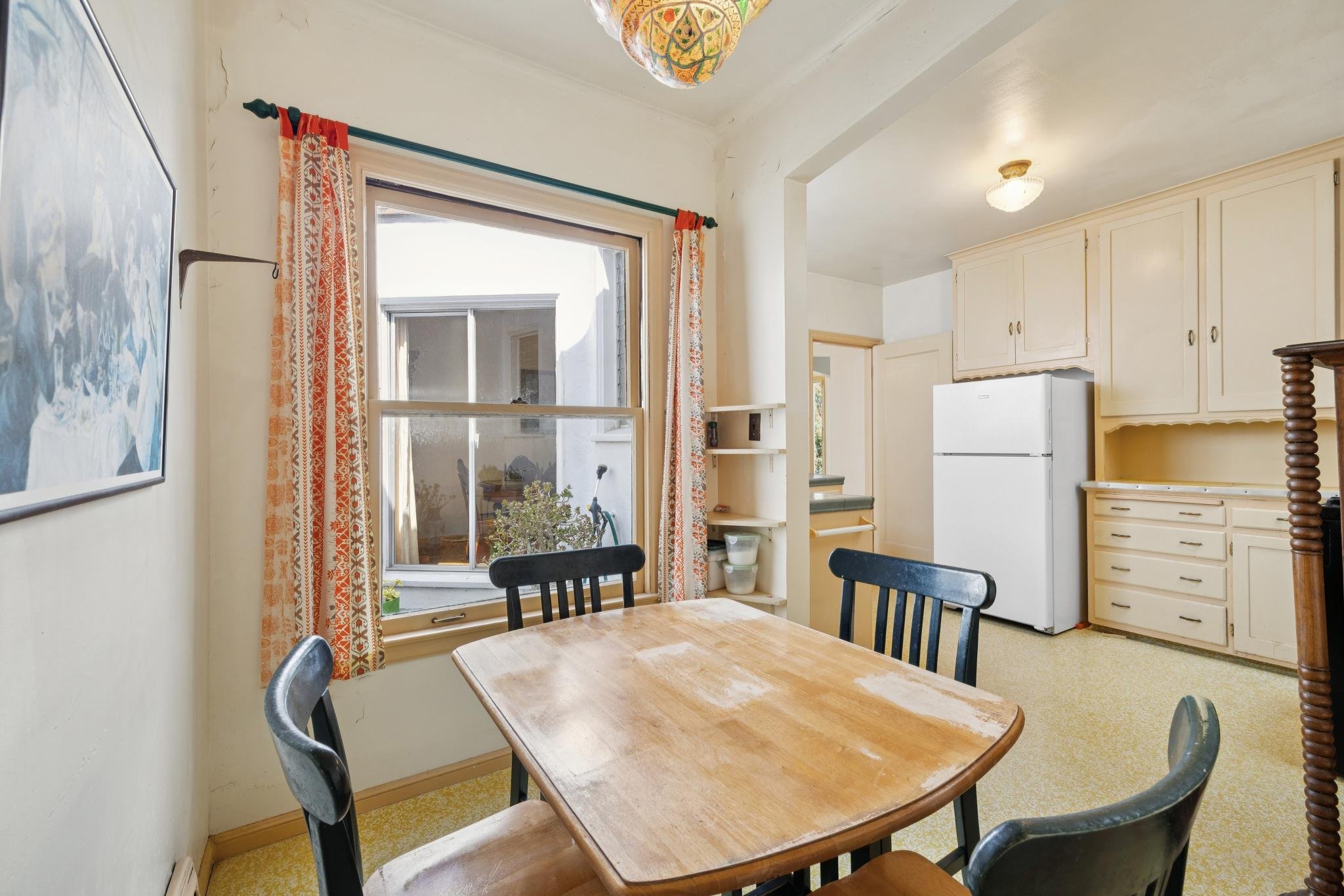 A view of a cozy kitchen and dining area with a wooden table, black chairs, a window with colorful curtains, and white cabinets with a refrigerator. There is a decorative hanging lamp on the ceiling and a picture on the left wall.