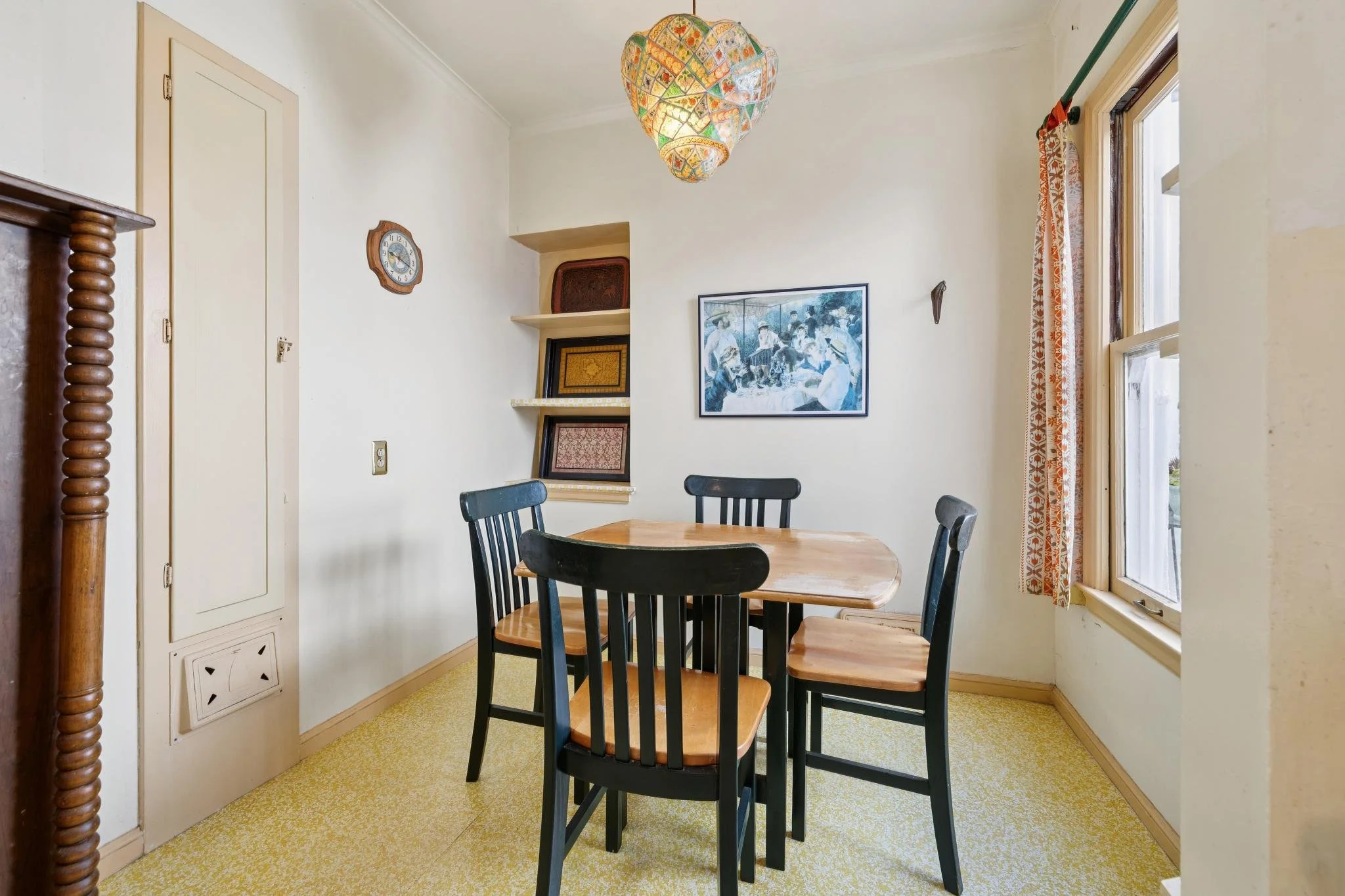 A dining area with a round wooden table surrounded by five black chairs, a window with orange patterned curtains, a white wall with a painting of people, a wall clock, and shelves with framed art, in a room with yellow carpet and a colorful hanging l