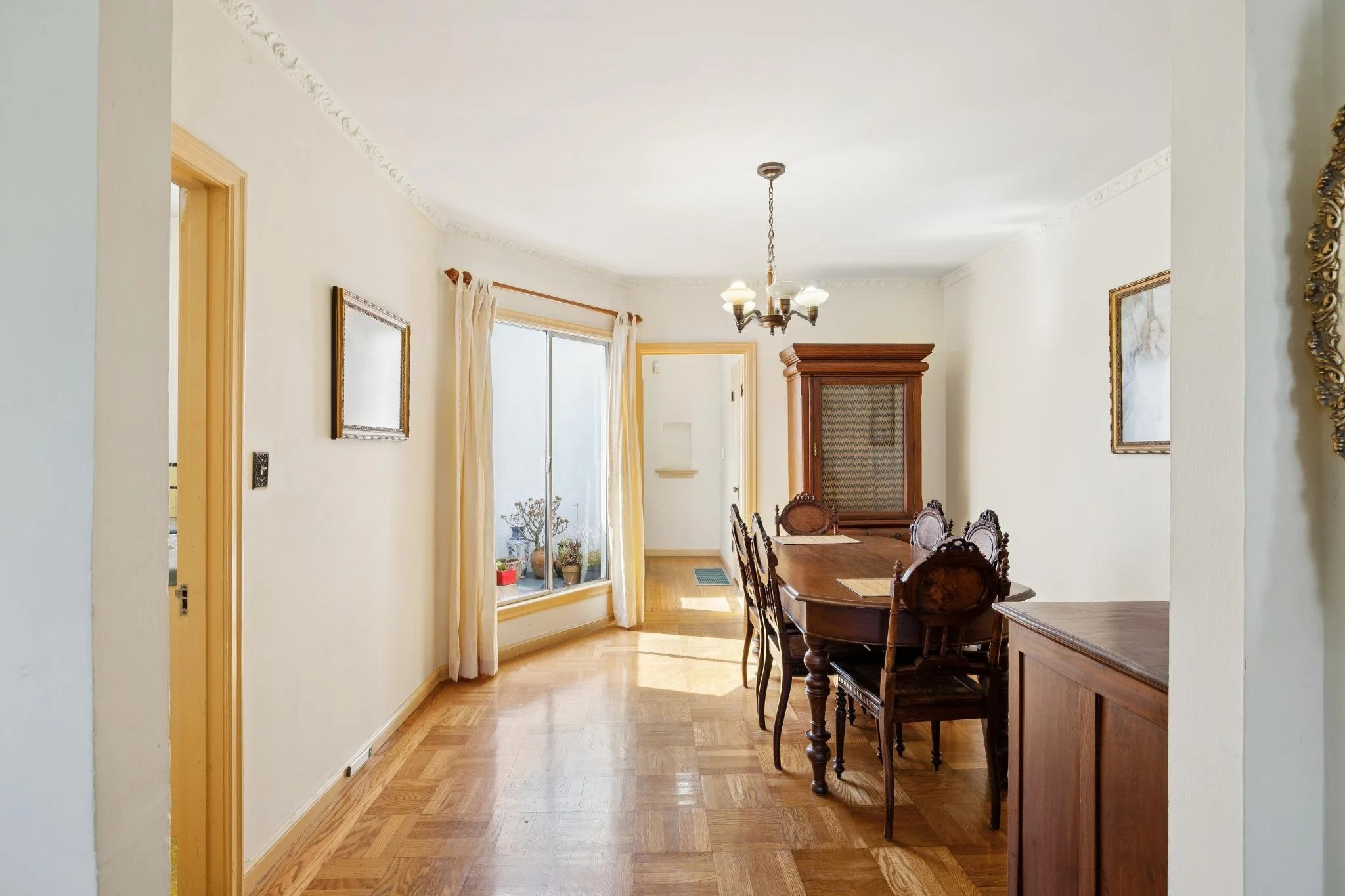 Dining room with wooden table and chairs, off-white walls with crown molding, large window with cream curtains, hardwood floors, and a vintage chandelier hanging from the ceiling.