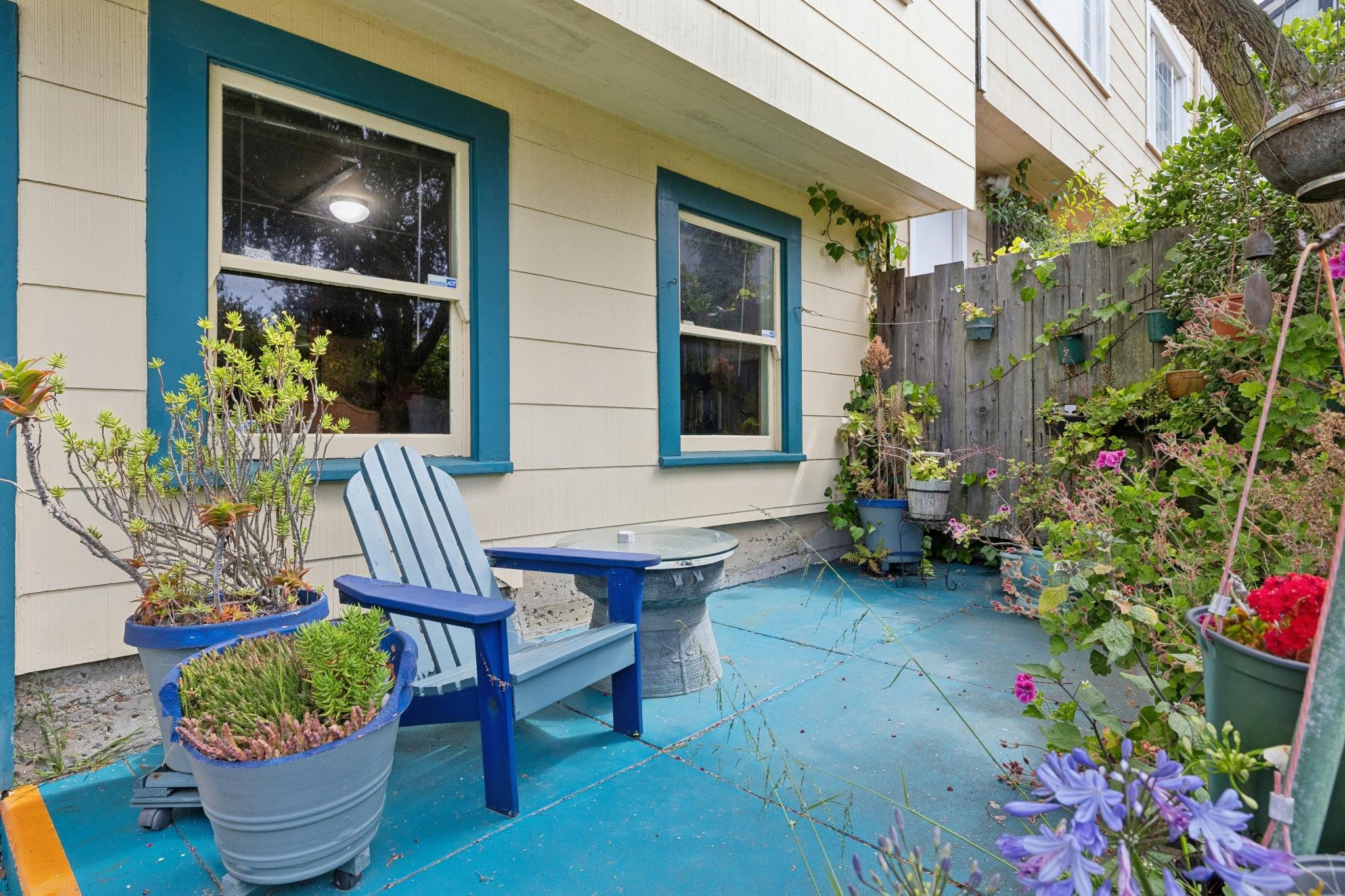 A cozy outdoor patio with potted plants, colorful flowers, and a blue wooden chair next to a small round table, adjacent to a house with yellow siding and two windows.