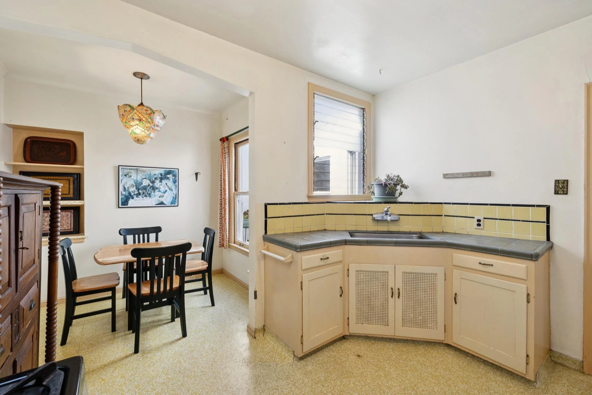 Interior view of a kitchen and dining area with a corner yellow tiled counter, a window with a plant on the sill, a small dining table with four black chairs, decorative pictures on the wall, and a hanging lamp with colorful stained glass design.