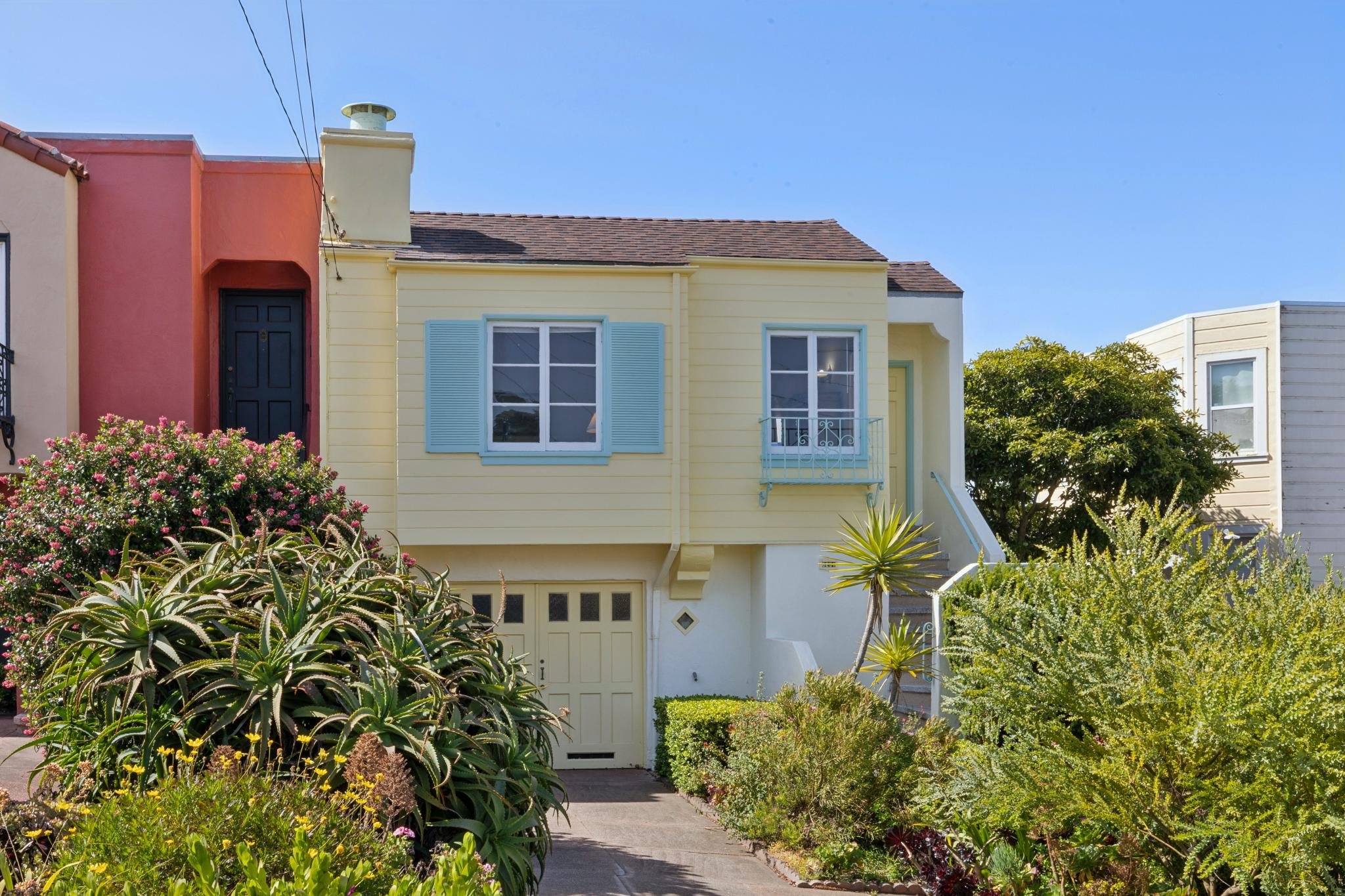 Colorful house with yellow siding, blue window shutters, and a black door, surrounded by lush greenery and flowers under a clear blue sky.