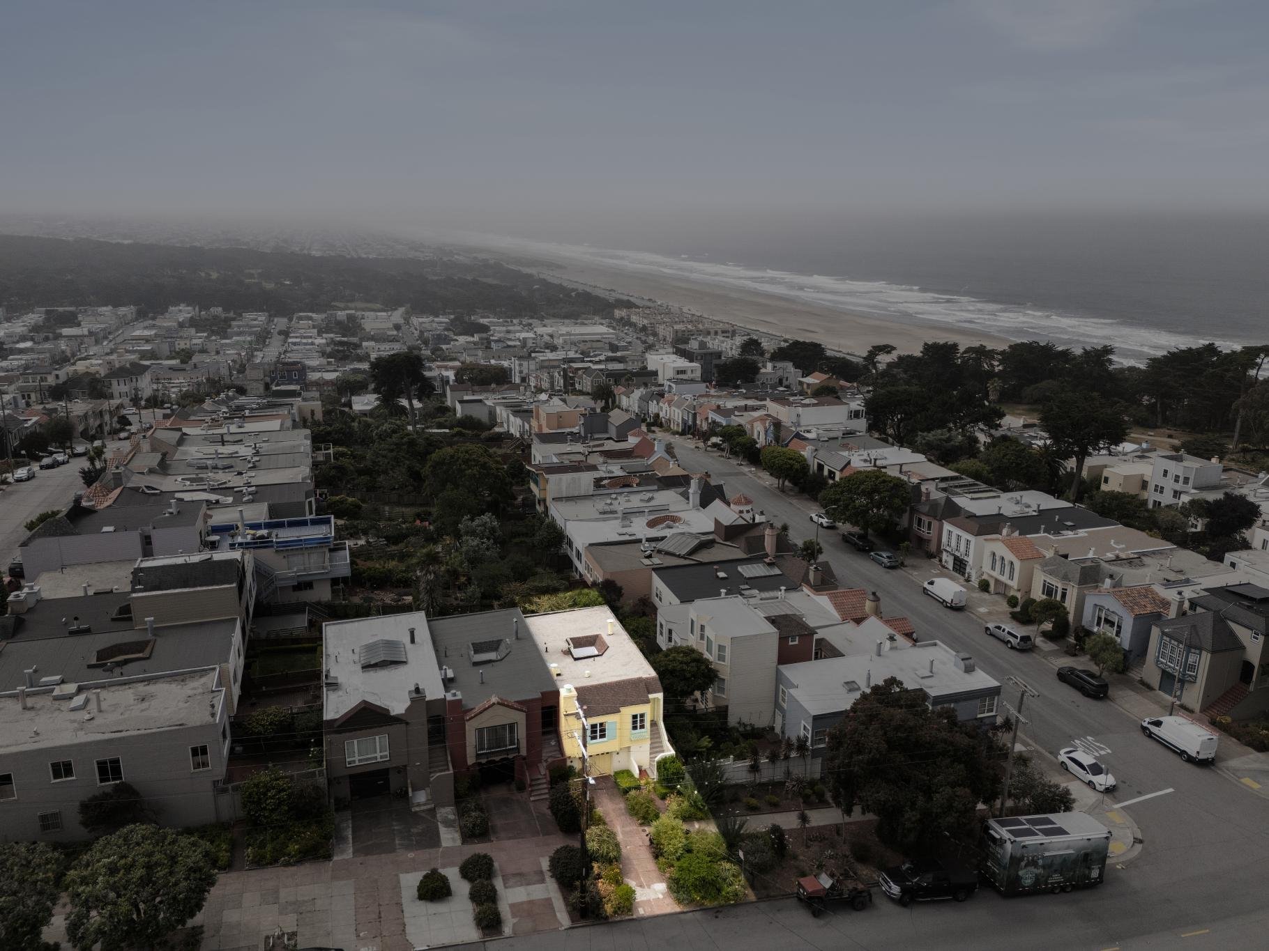 A coastal cityscape with houses and streets in the foreground, and a beach with ocean waves fading into the horizon under a cloudy sky in the background.