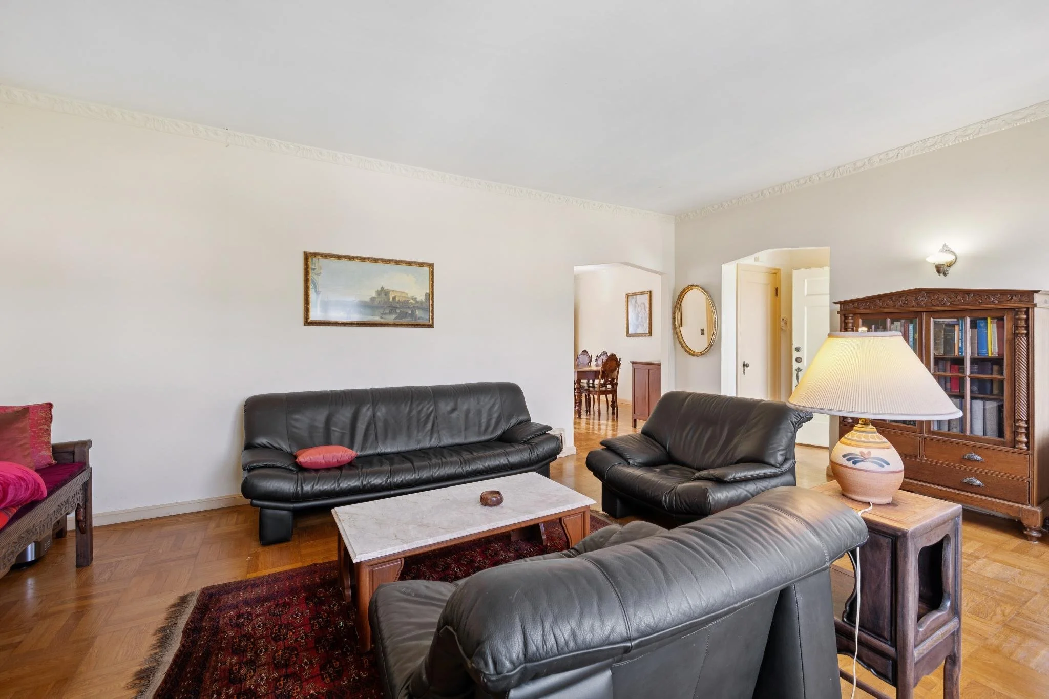 A living room with black leather sofas, a marble coffee table, a patterned area rug, a wooden bookshelf, a table lamp, and framed artwork on white walls and hardwood flooring.