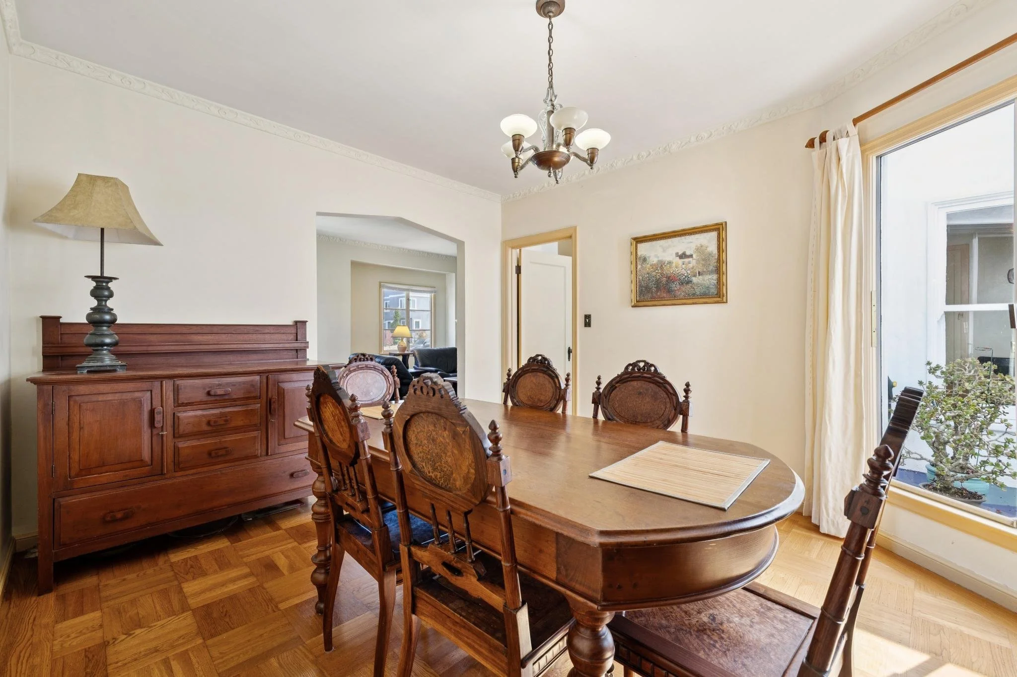 A dining room with a large wooden table and six matching chairs, a wooden sideboard with a table lamp, a chandelier hanging from the ceiling, a painting on the wall, large windows with white curtains, and a view of a neighboring house and greenery ou