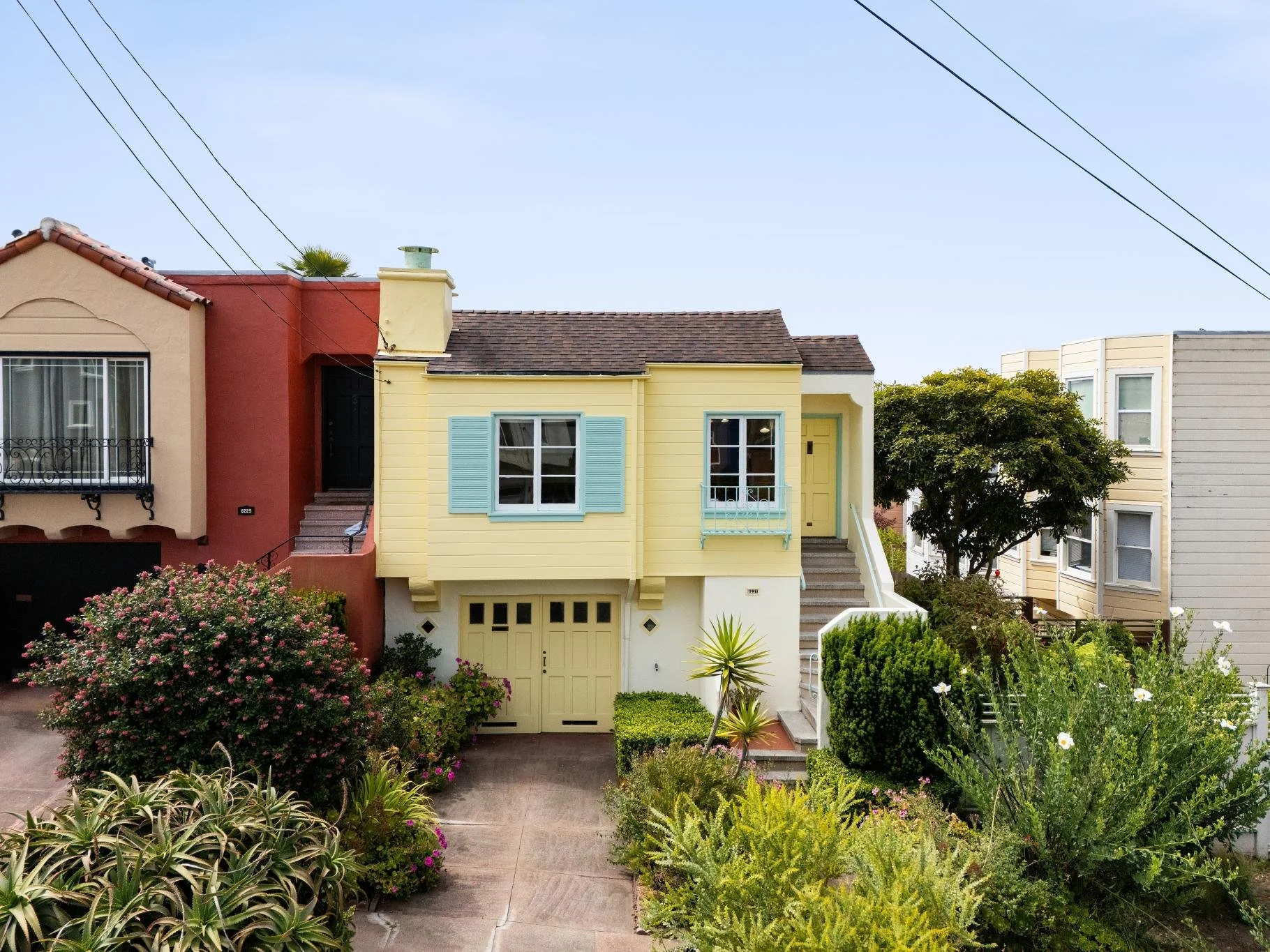 Colorful residential houses with front gardens, bushes, and flowers in a neighborhood under a partly cloudy sky.