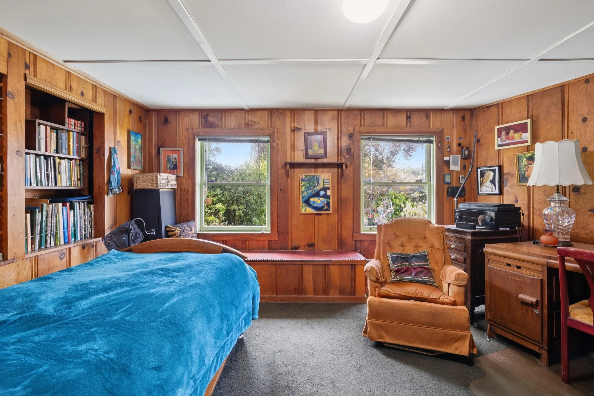A cozy living room with wooden paneled walls, two large windows showing greenery outside, a bed with a blue cover, a comfortable orange armchair, a wooden media console with a speaker system, many framed artworks on the walls, and a table lamp.