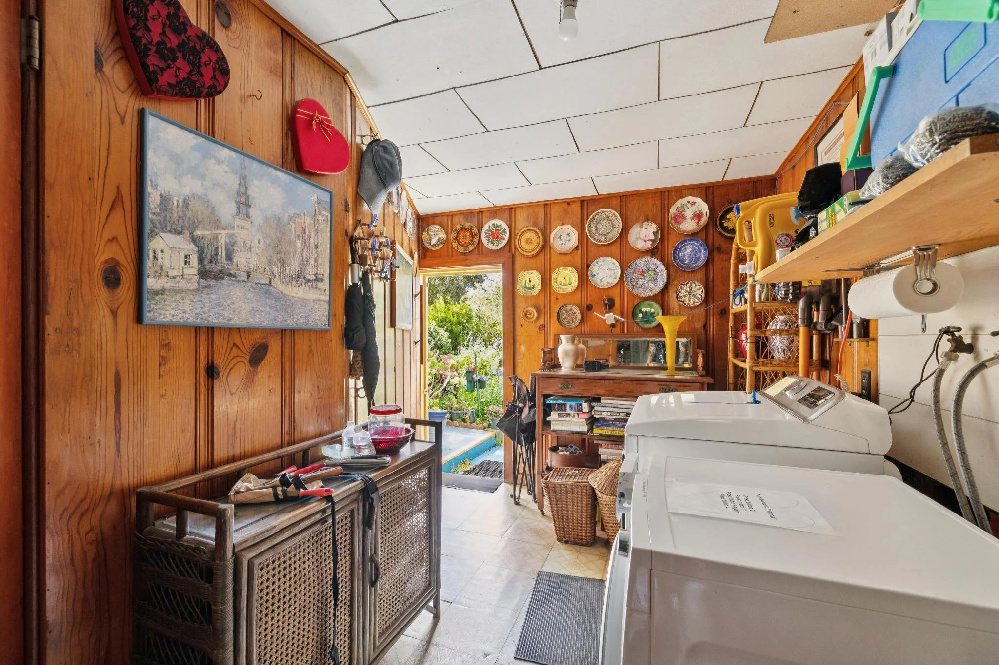 A laundry room with wood-paneled walls decorated with painted plates. There are laundry supplies, a washing machine, and a laundry basket. The room opens to a garden outside.