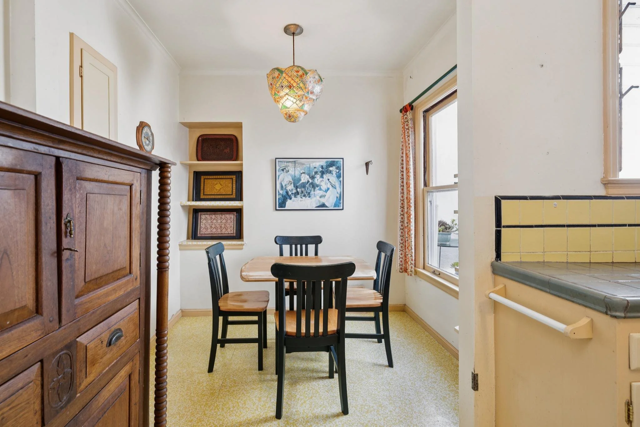 A dining area with a wooden table and four black chairs, a vintage wooden cabinet, framed artwork on the wall, and a colorful hanging light fixture. There are windows with curtains allowing natural light, and a tiled section of the kitchen is visible