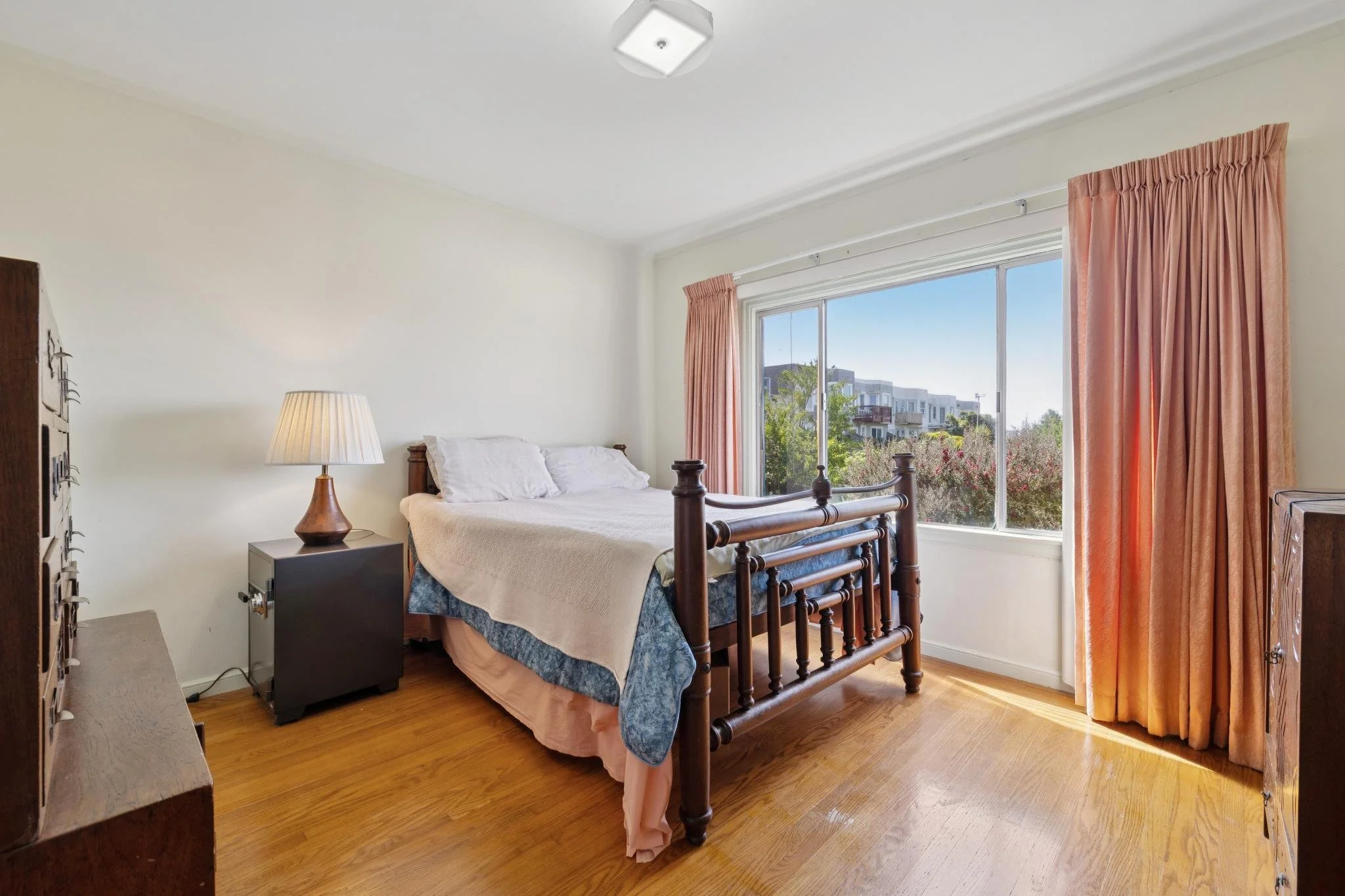 A bedroom with a wooden bed, white bedding, a nightstand with a lamp, large window with pink curtains, and a view of modern buildings outside.