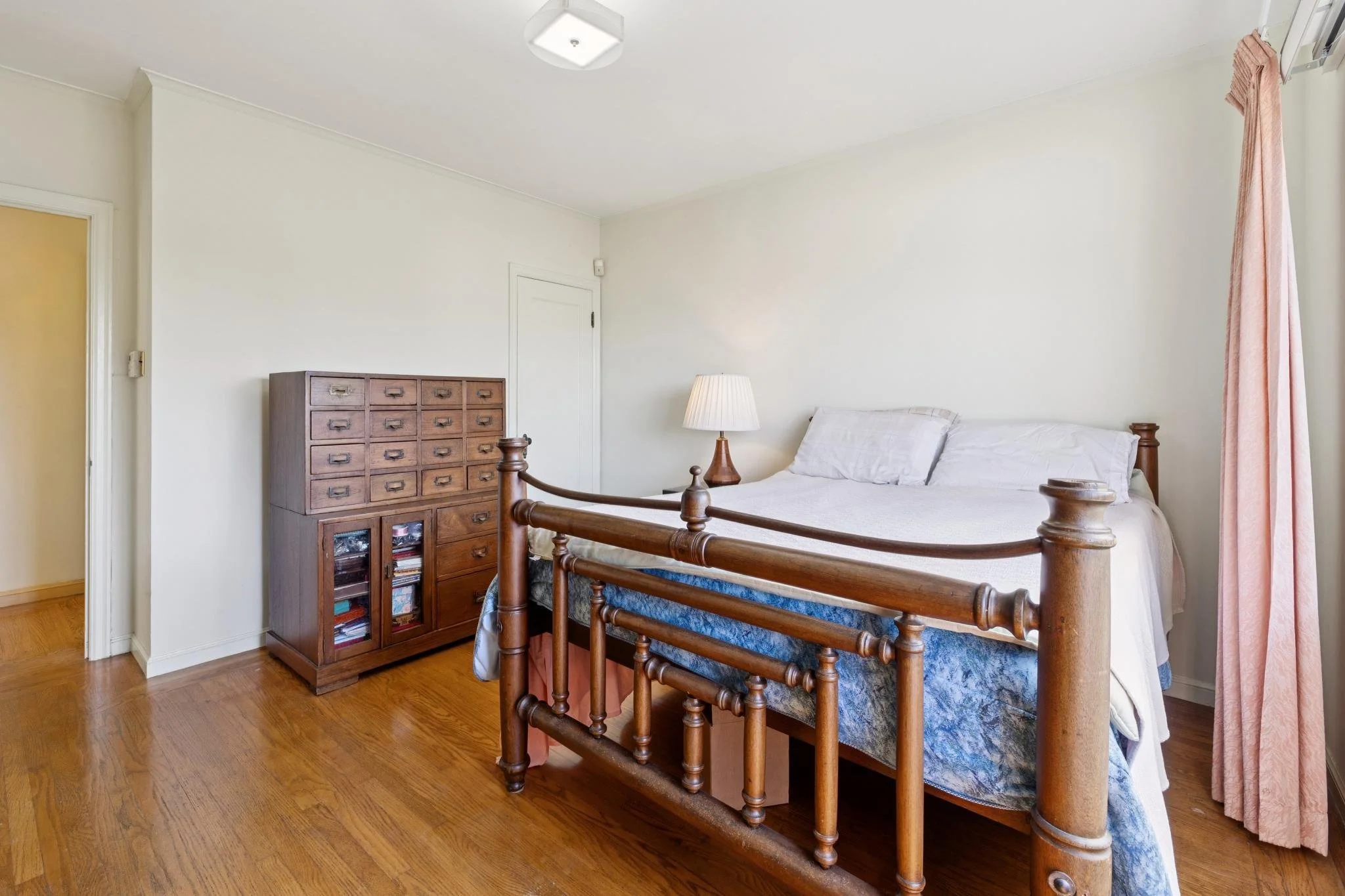A bedroom with a wooden bed, nightstand, and a lamp on it, white walls, a window with pink curtains, and a wooden dresser, with hardwood floors.