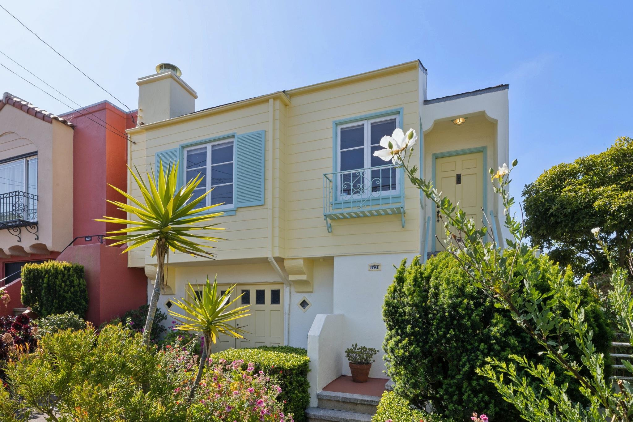 A yellow two-story house with blue shutters and a small balcony on the upper floor, surrounded by green bushes, flowering plants, and trees, under a clear blue sky.