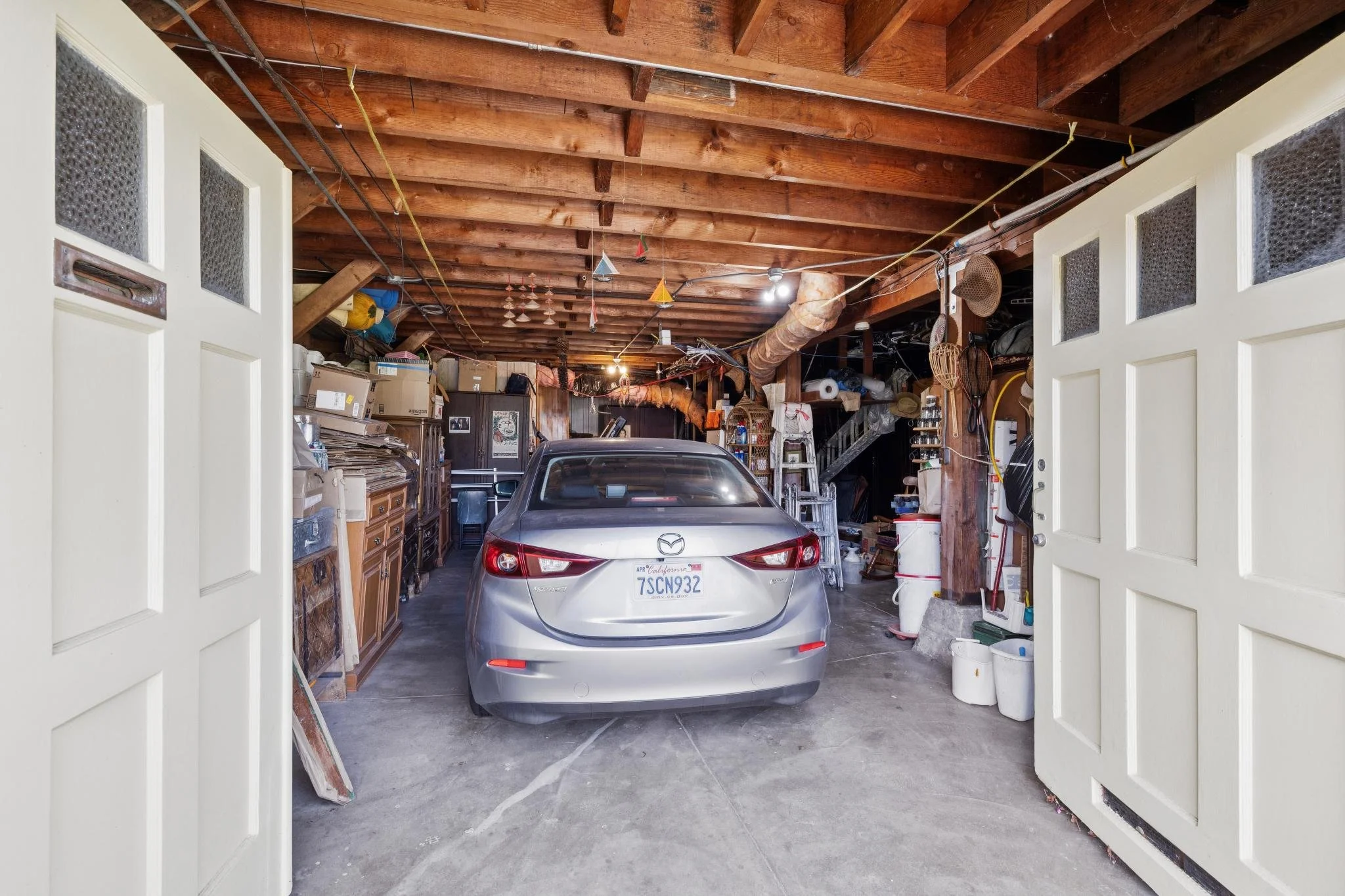 View of a garage filled with miscellaneous items, a silver Mazda car parked inside, and a wooden ceiling with exposed beams and hanging decorations. Double doors open into the garage.