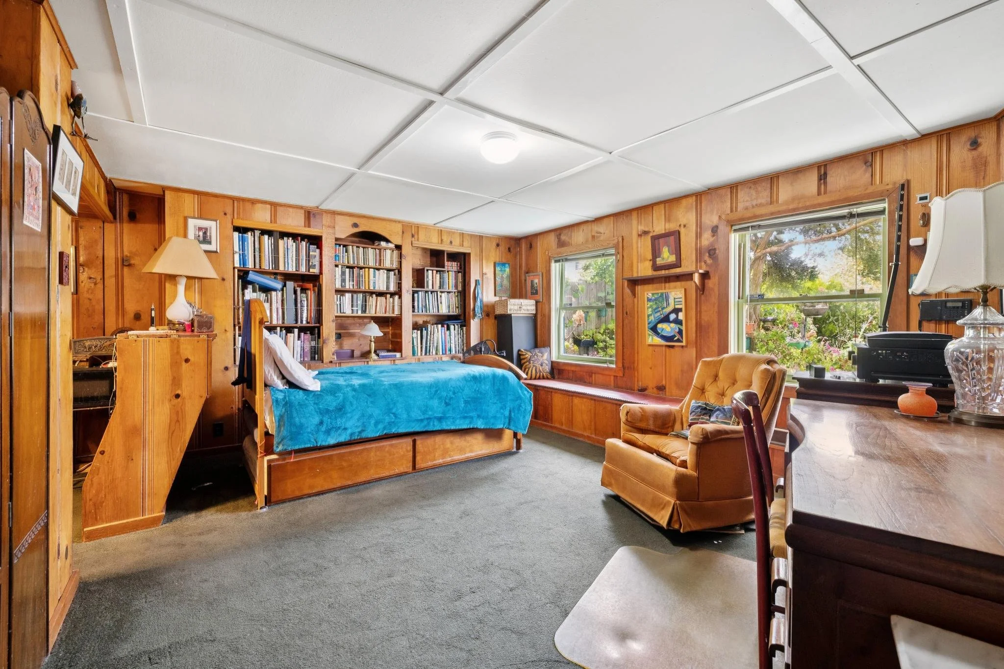 Living room with wooden walls, bookshelves filled with books, a bed with a blue blanket, windows showing greenery outside, and furniture including a chair, table, and lamp.
