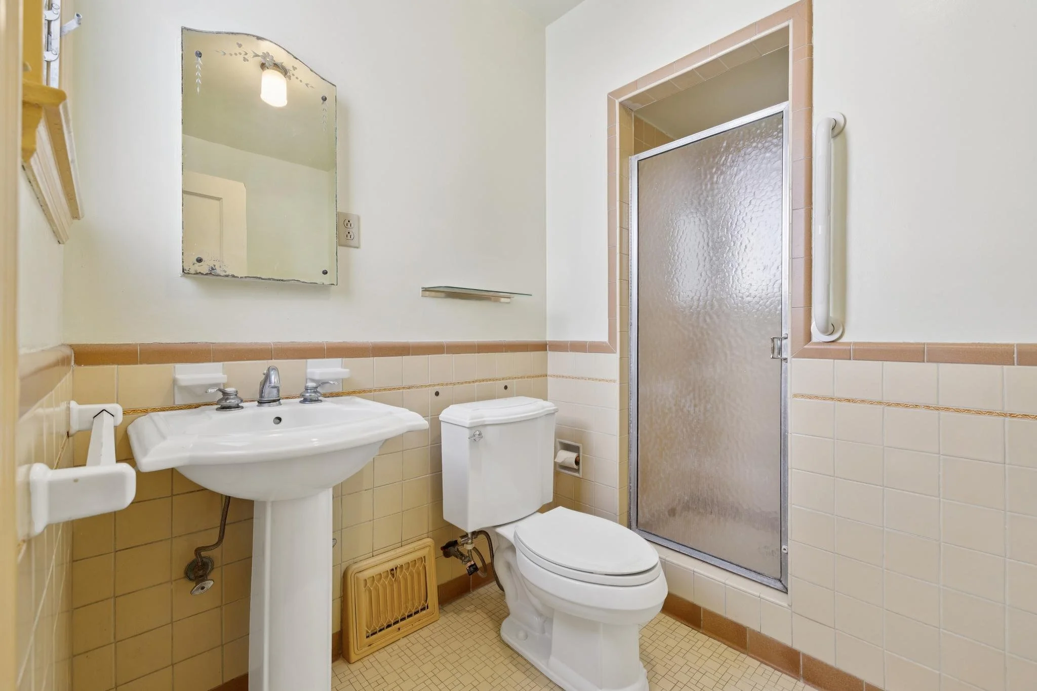 Small bathroom with yellow tile on the lower walls and cream paint on the upper walls. Features a white sink with a silver faucet, a toilet, a rectangular mirror, a shower with textured glass door, and a wall-mounted grab bar. The fixtures are standa