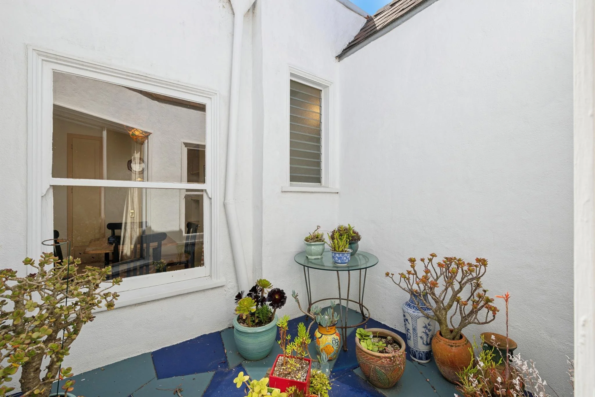 A small outdoor patio with various potted plants and succulents near a white house wall with a window and a glass top table.