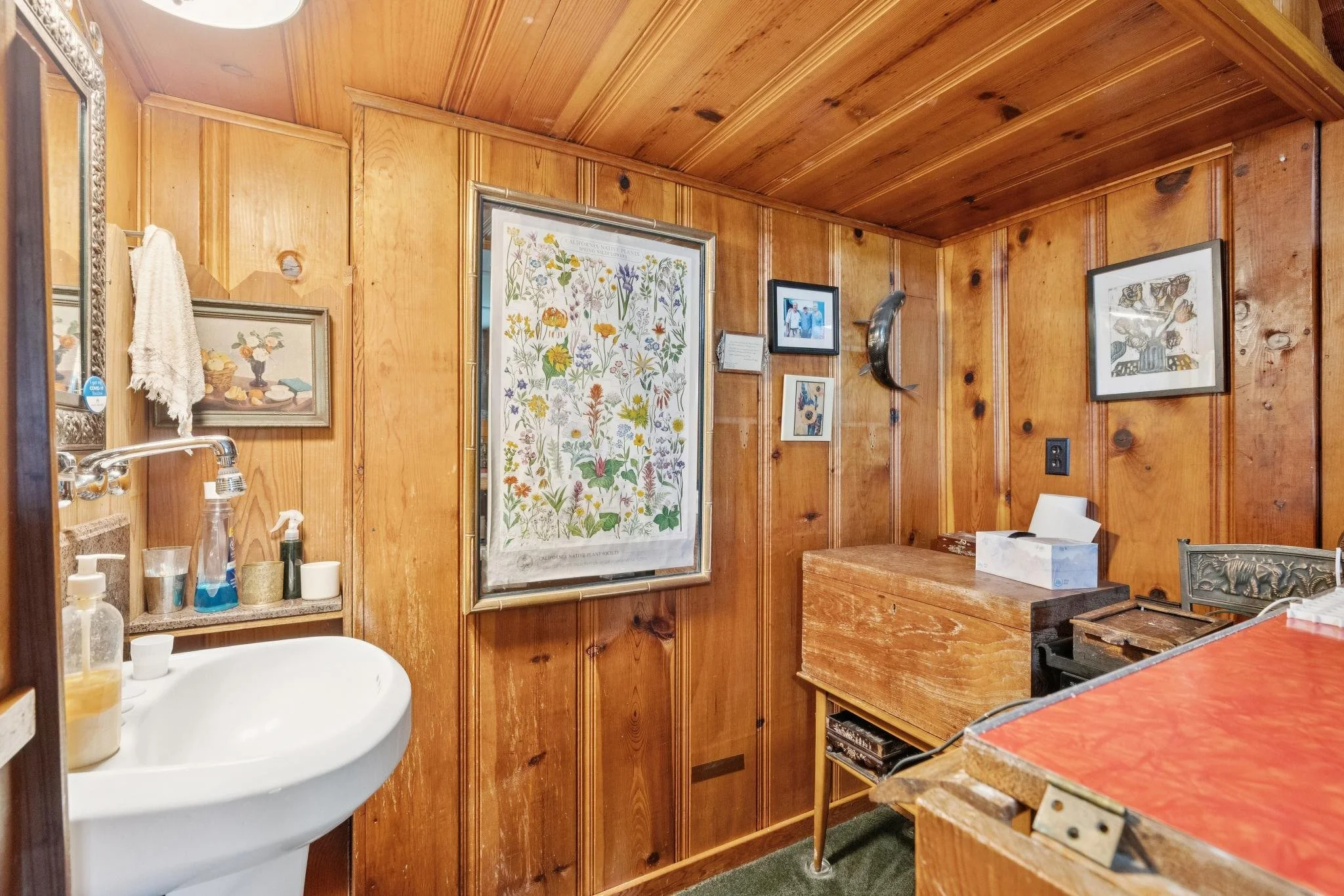 Interior view of a wooden-paneled bathroom with a sink, various toiletries, framed botanical art, and additional framed pictures and decorations on the walls.