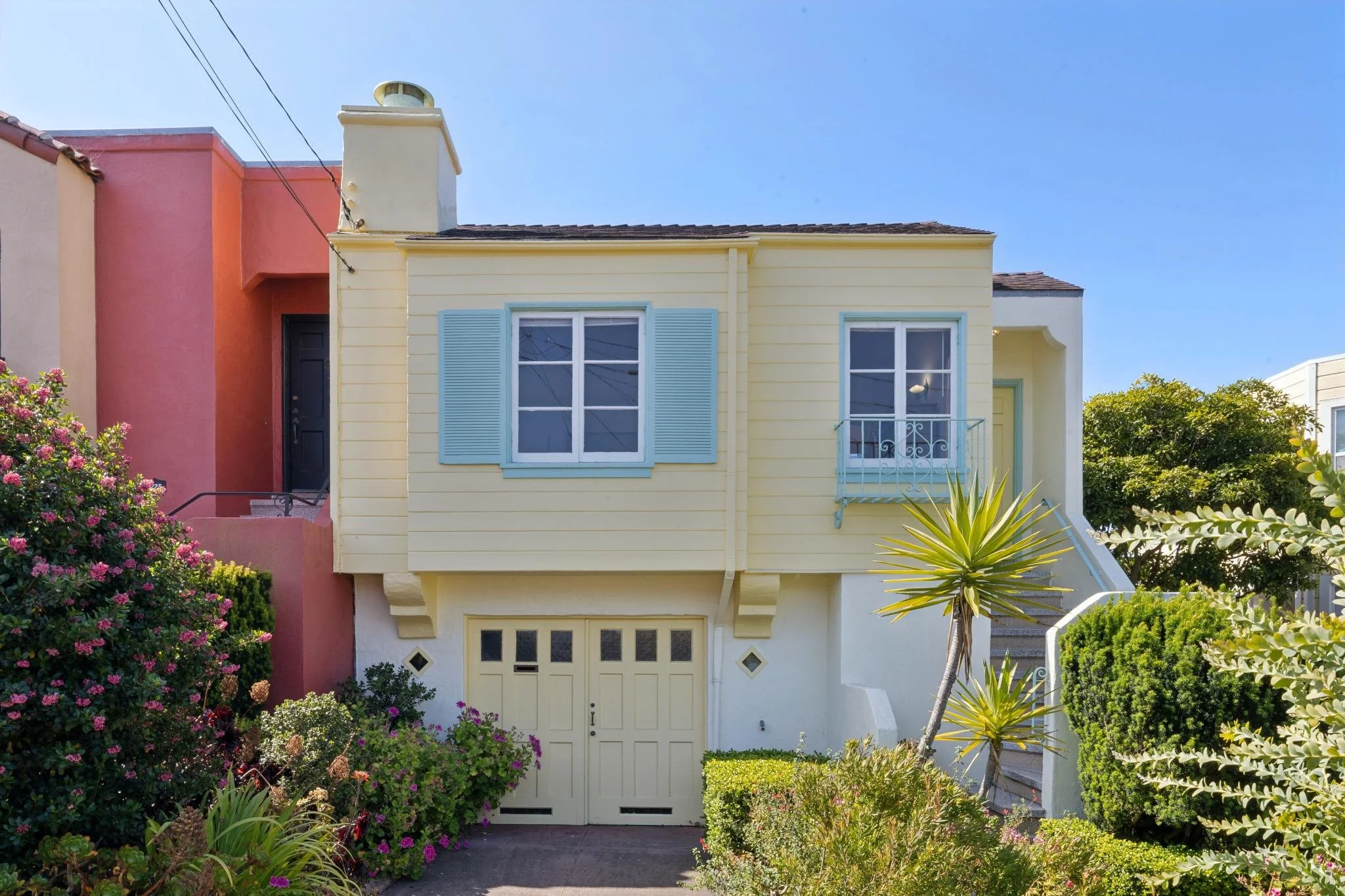 Colorful house with a yellow facade, white window shutters, and a small balcony, surrounded by lush green bushes, flowering plants, and a clear blue sky.