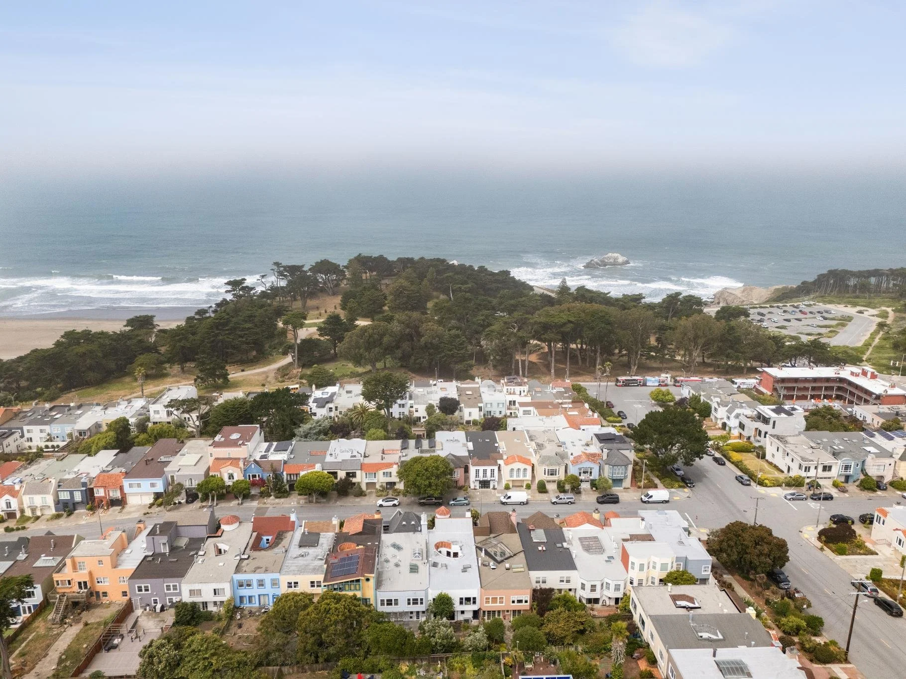 An aerial view of a coastal neighborhood with colorful houses, a park with trees, and a beach with waves in the background.