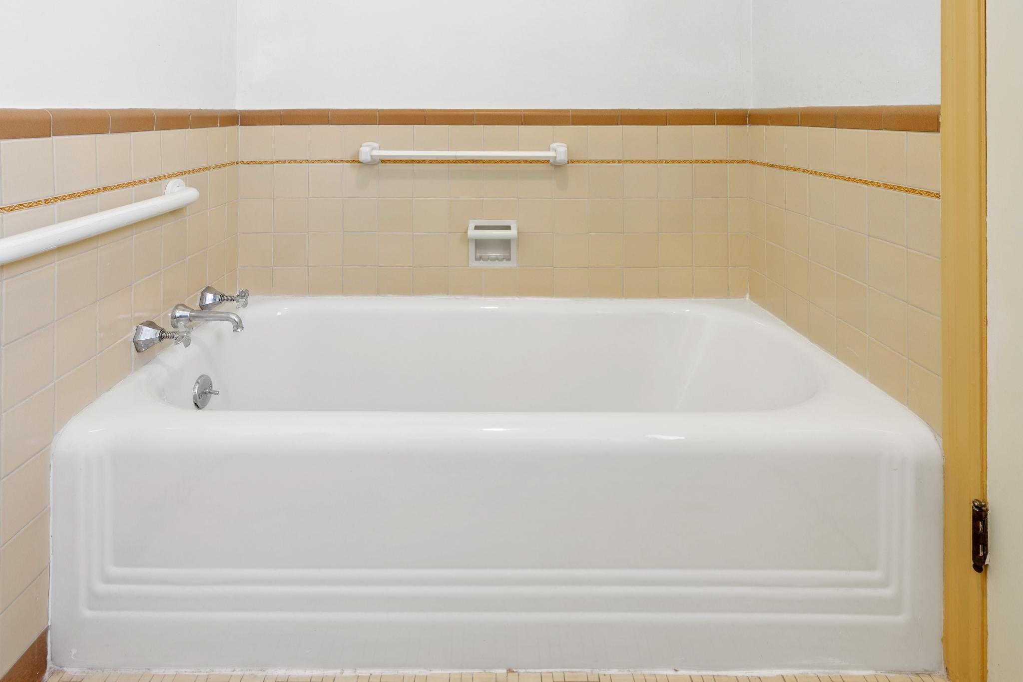 Shower or bathtub area with tan tiled walls, white grab bars, soap dish, and a yellow door frame.
