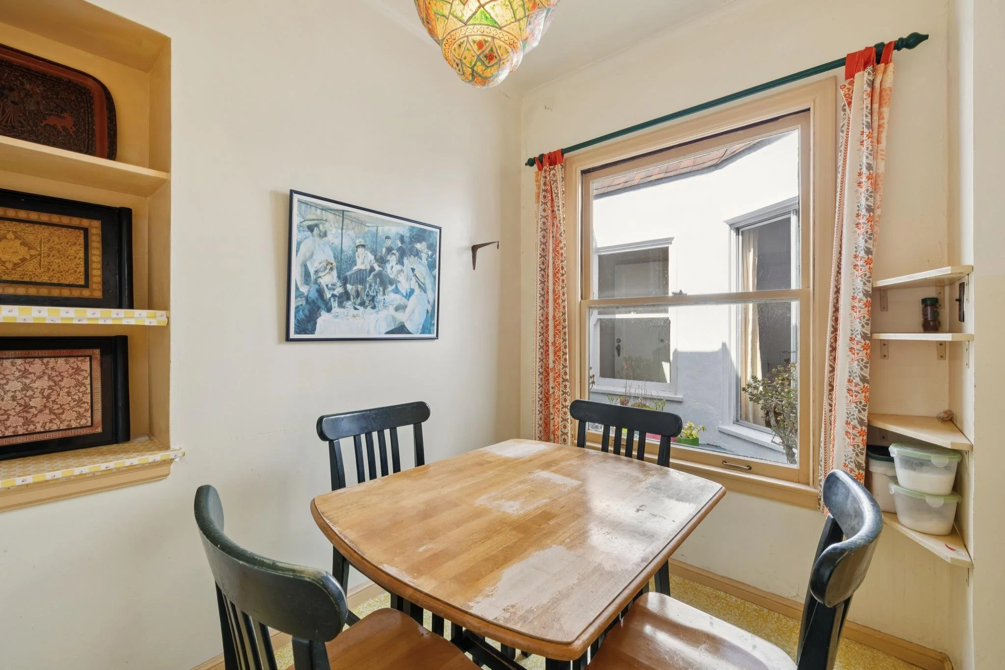 A cozy kitchen corner featuring a wooden dining table with four black chairs, a window with floral curtains, a colorful hanging lamp, a small wall shelf, and decorative artwork on the wall.