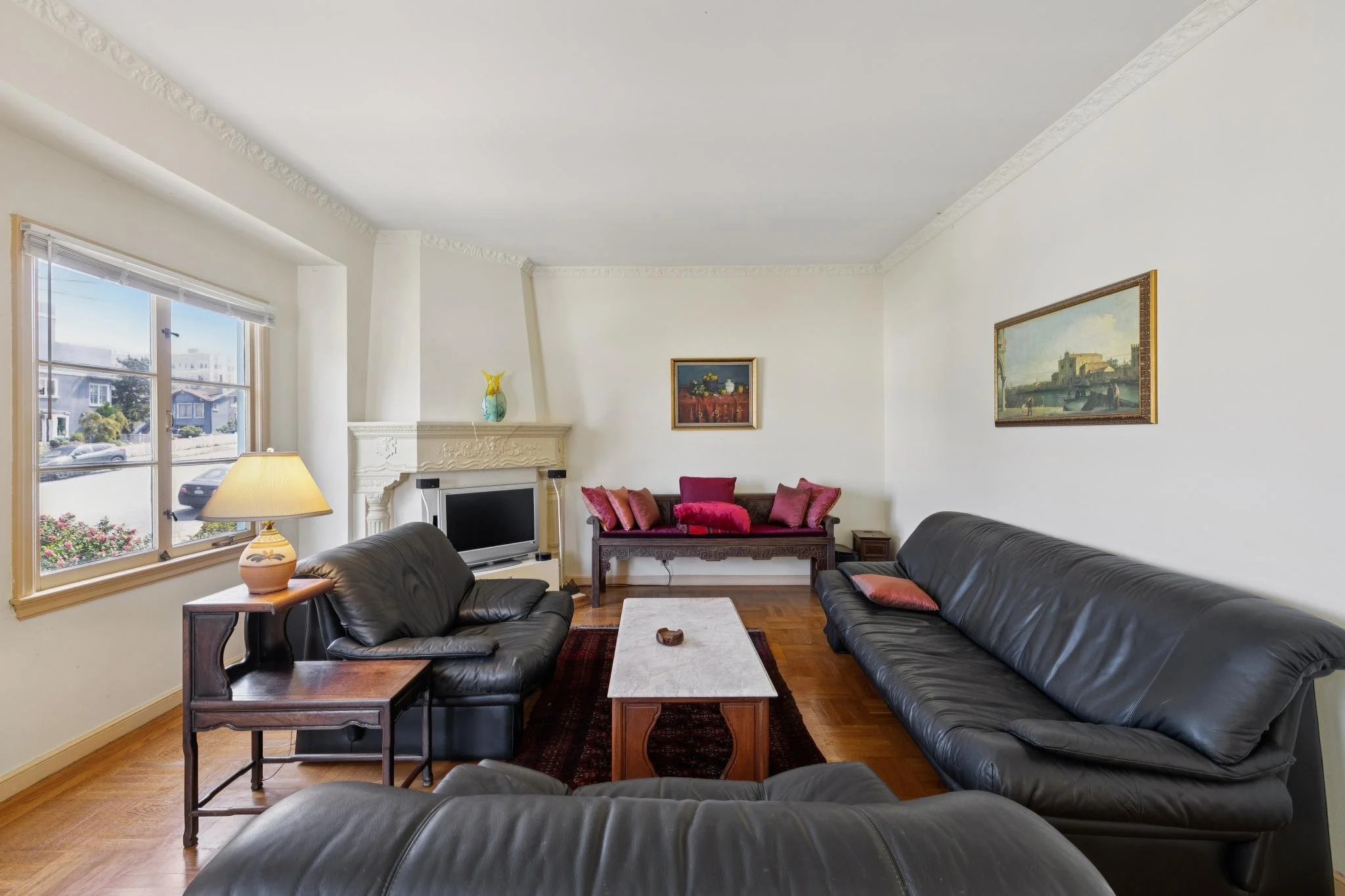 A living room with large windows on the left, black leather sofas, a wooden coffee table, and framed artwork on the walls.