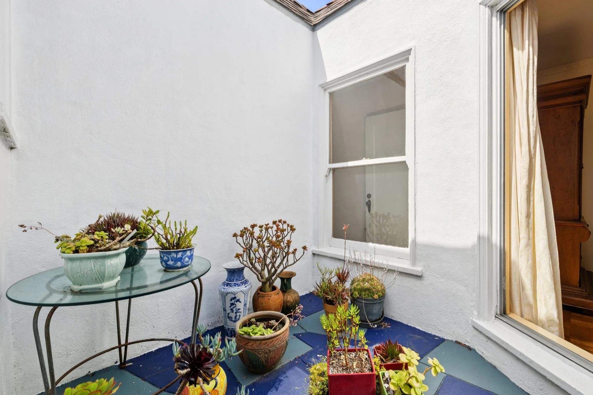 A small balcony with a glass table surrounded by various potted plants, including succulents, on a blue-tiled floor, with a white exterior wall, a window, and a door with a curtain in the background.