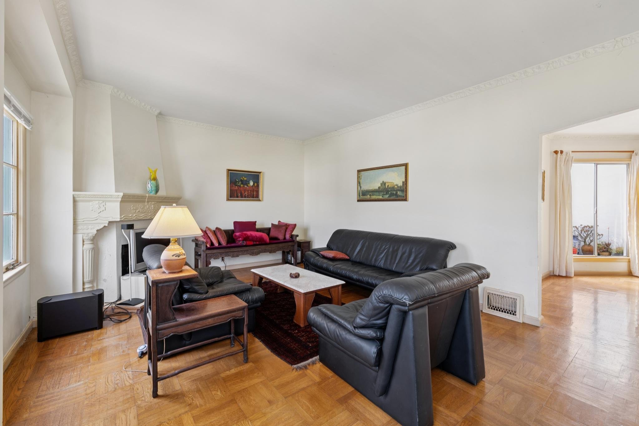 Living room with black leather sofa and wooden furniture, paintings on white walls, hardwood floors, window with curtains, fireplace with a vase on mantle, lamp on side table, and two speakers on floor.