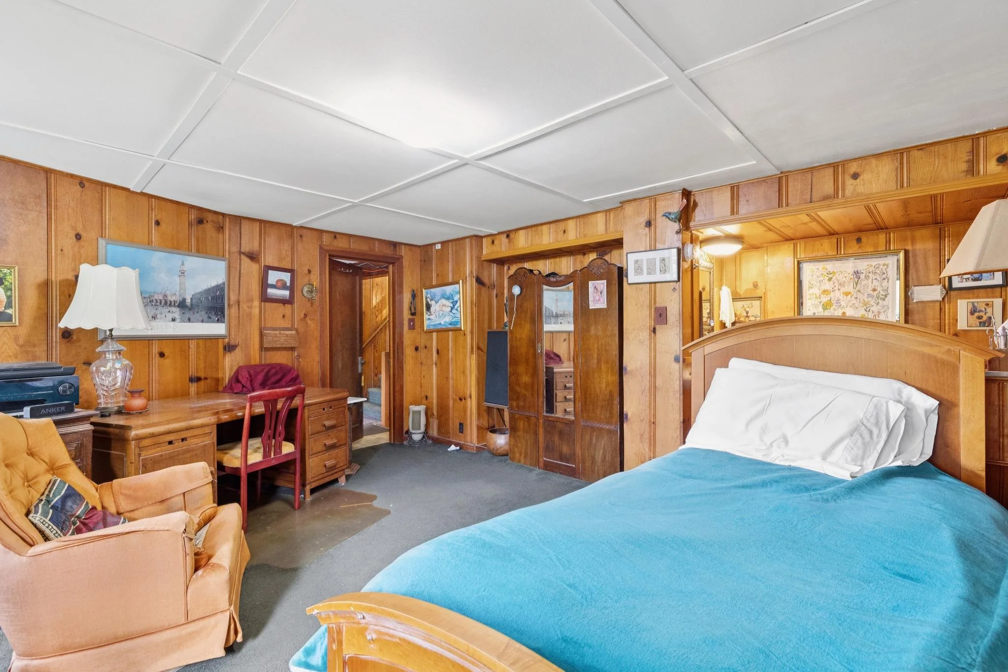 Interior of a bedroom with wood-paneled walls, a bed with turquoise bedding, an orange armchair, a wooden desk with a lamp and electronic devices, and framed artwork on the walls.