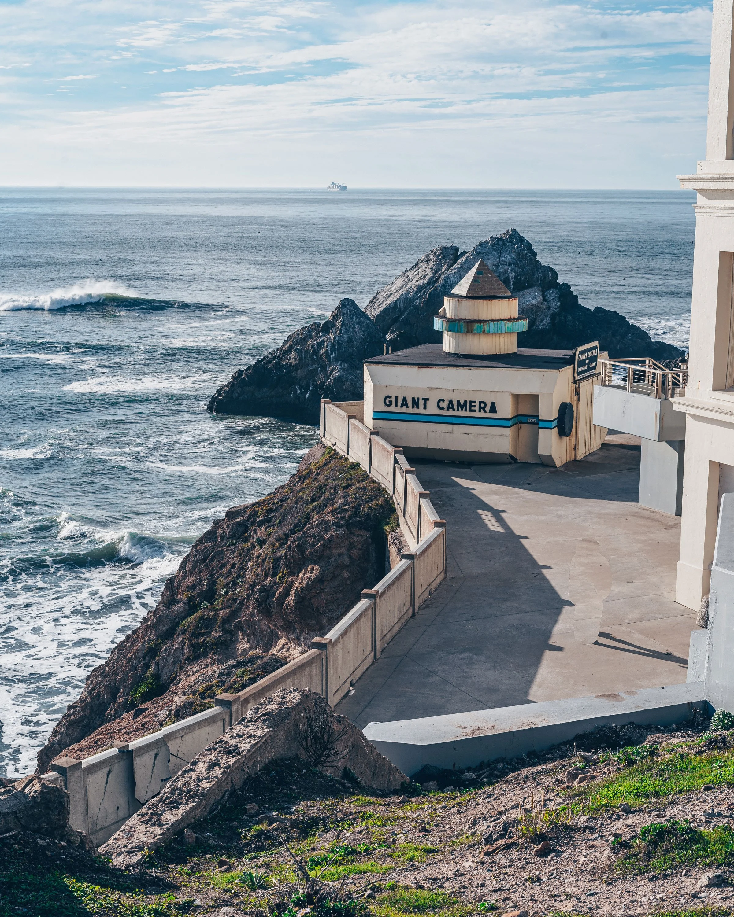 View of the ocean with waves hitting rocks along the coast, a small building with the sign 'Giant Camera', and a distant ship at sea under a partly cloudy sky.