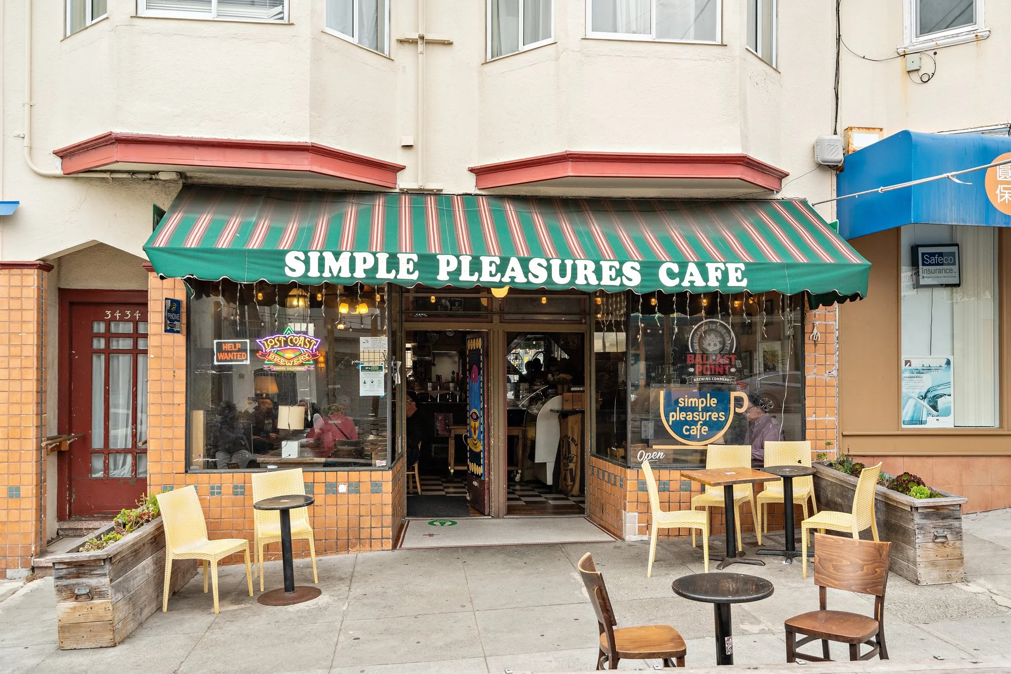 The image shows the exterior of a café named Simple Pleasures Café, with an awning and window seating. There are several chairs outside, a few small tables, and a brick and tile facade. Inside, visible through the large window, are people eating, and