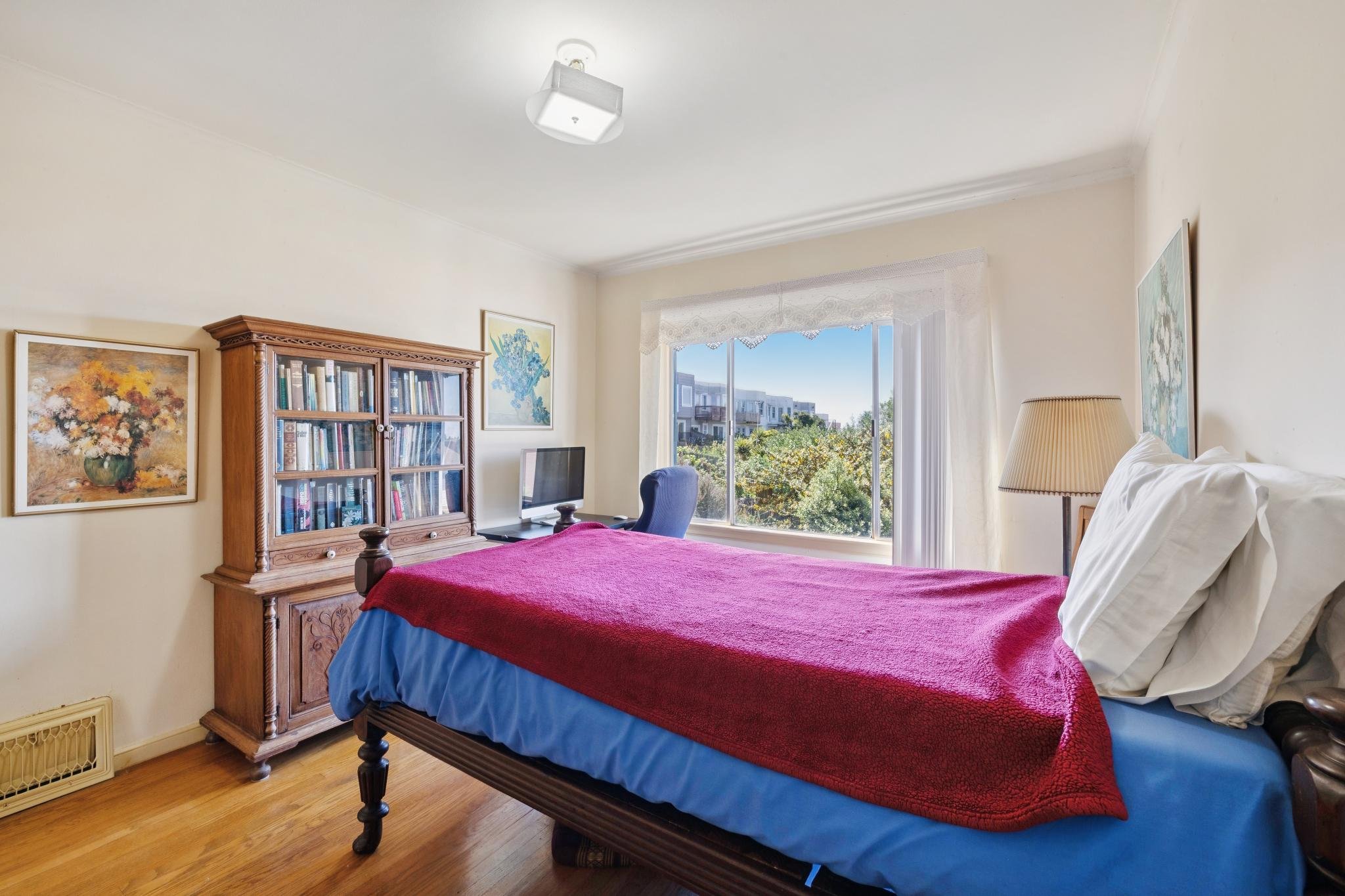 A bedroom with a large window showing trees and a building outside. The bed has a red blanket and white pillows. There is a wooden bookshelf with books and art on the walls. A small desk with a computer and a blue chair is near the window. A lamp is 