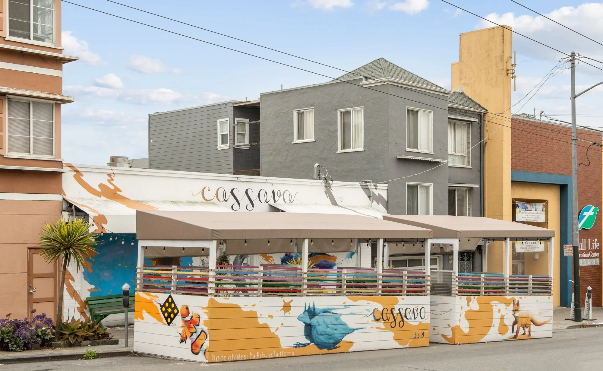 Colorful mural on a fence featuring a blue globe, butterflies, and abstract art. Background shows multi-story buildings, utility lines, and a partly cloudy sky.