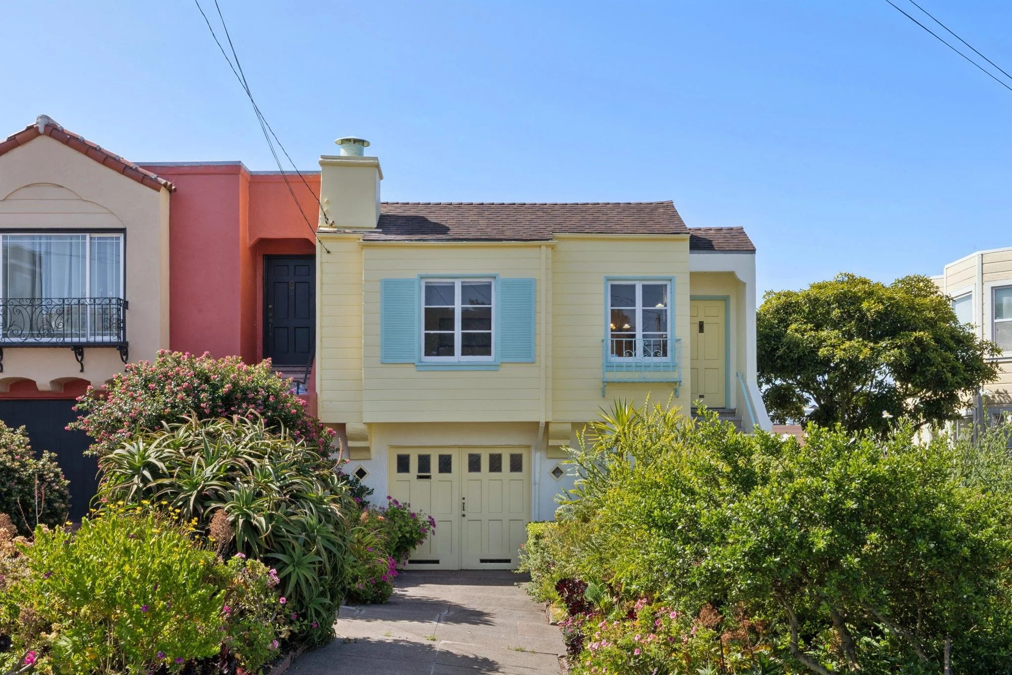 Front view of a colorful row of houses with garden plants and trees in the foreground under a blue sky.