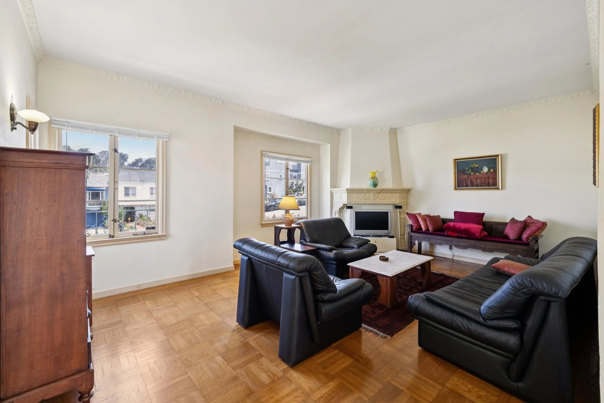 Living room with multiple black leather sofas, wooden coffee table, and a fireplace with decorative molding. Large windows with views of neighboring houses and trees, and a painting on the wall.