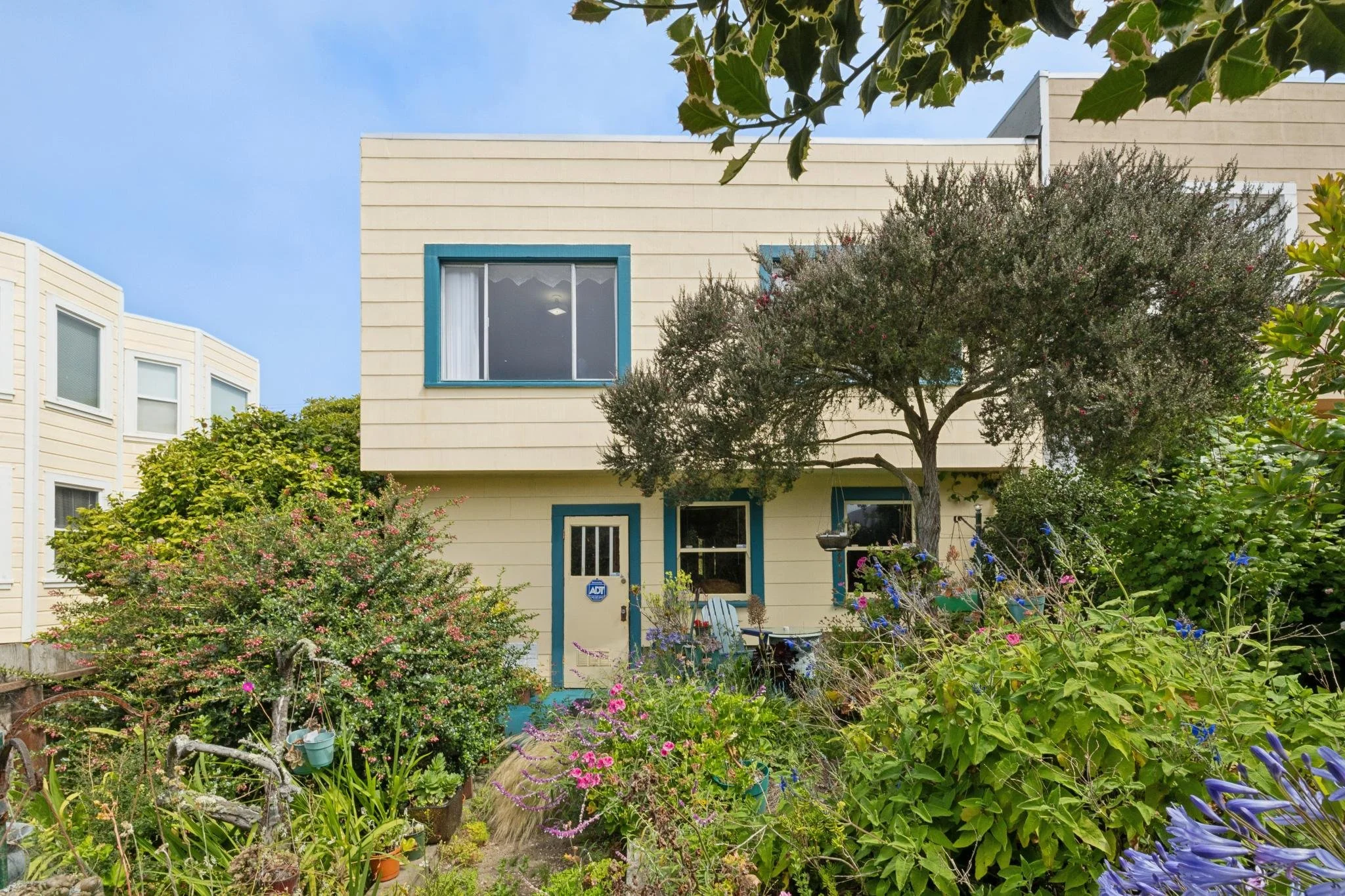 A yellow house with blue trim surrounded by a lush garden with various plants, flowers, and trees.