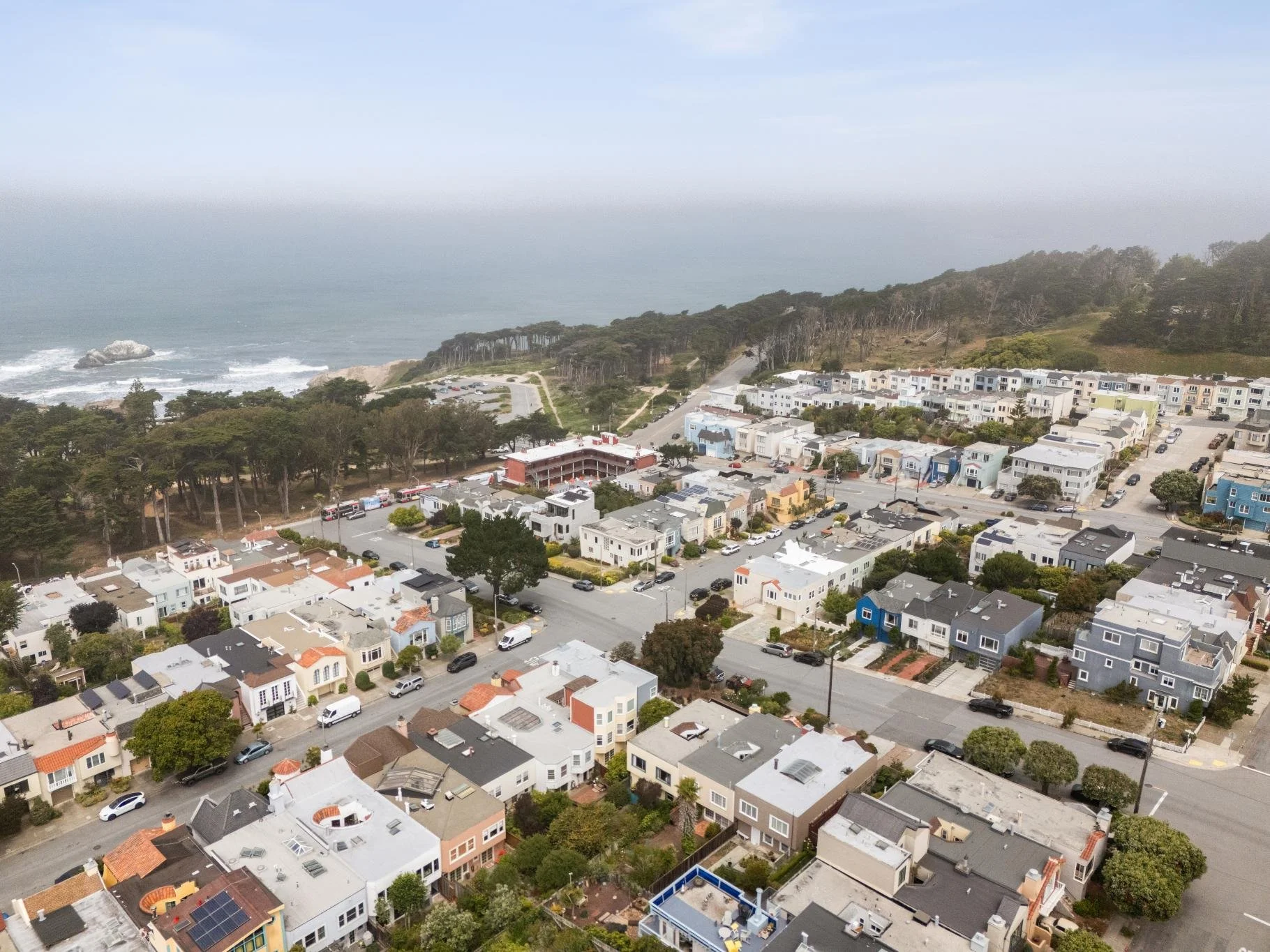 An aerial view of a coastal neighborhood with a mix of houses and apartment buildings, trees lining the streets, and the ocean in the background with waves crashing against rocks.