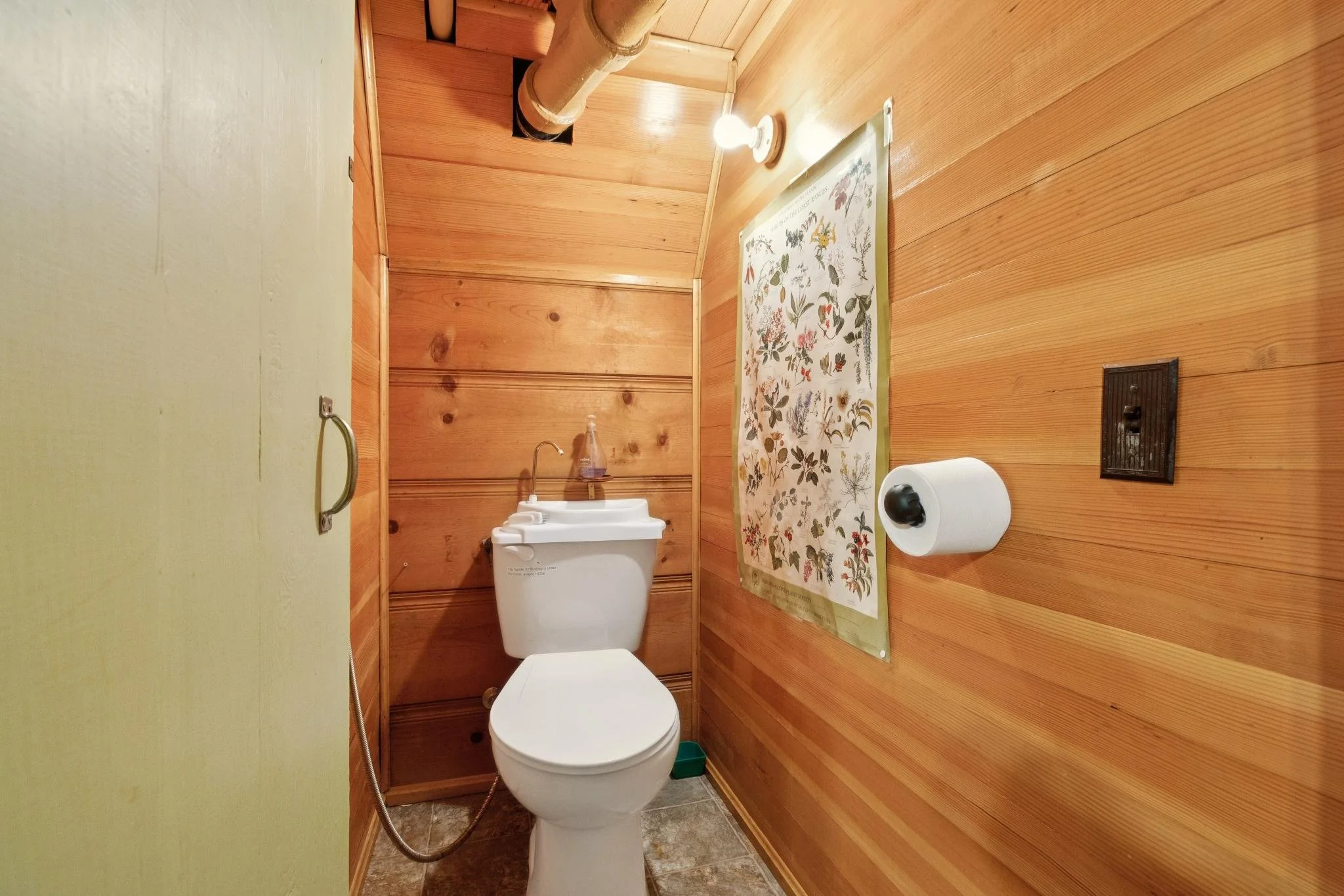 A small bathroom with wood-paneled walls, featuring a white toilet, a small sink with a bottle of soap, a toilet paper roll, a vent, a framed botanical print, and a wall-mounted black electrical switch.